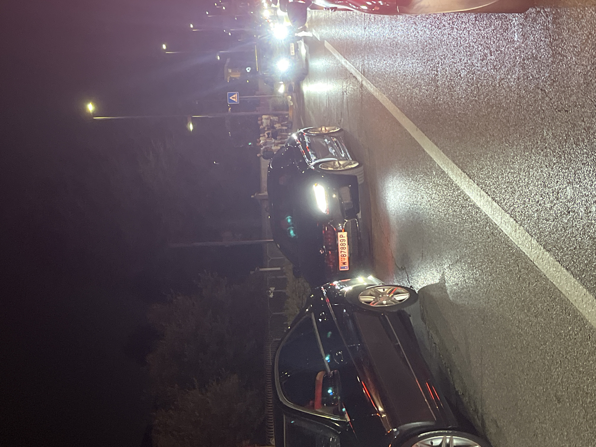 The image captures a nighttime street scene in Castiglione delle Stiviere, Italy, likely a road with streetlights lining the side. Several cars are present, with at least two dark sedans prominently featured. One car, with a license plate reading "W-87889-P," appears to be a sporty model with gold rims and illuminated headlights. The road surface is wet, reflecting the light from the vehicles and street lamps, indicating recent rain or damp conditions. In the background, a group of people are gathered on the sidewalk, some appearing to be looking at the cars. The overall atmosphere suggests a gathering or event occurring at night.