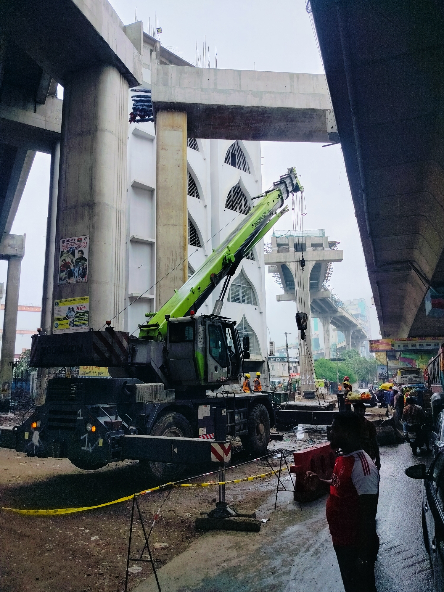 In Dhaka, Bangladesh, an active construction site is depicted. A large green crane, identified as a ZOOMLION model, is positioned prominently, its boom extended upwards and outwards. The crane is situated on a dirt and mud-covered ground, indicative of ongoing heavy construction.
Several individuals, dressed in construction attire including hard hats and safety vests, are visible near the crane and the immediate work area. One person in the foreground wears a red and white shirt, looking towards the left of the frame. Other construction workers are spread across the scene, some attending to the crane or the surrounding materials.
The background reveals the ongoing construction of an elevated structure, likely a bridge or expressway, with large concrete pillars and beams forming the framework. Adjacent to the construction, a white building with arched windows is visible, suggesting an urban setting. The overall environment appears overcast and wet, with puddles of water on the ground and a generally damp atmosphere. The scene is bustling with activity, typical of a large-scale infrastructure project in a densely populated city like Dhaka. Traffic and other urban elements can be seen in the distance, further contextualizing the location.