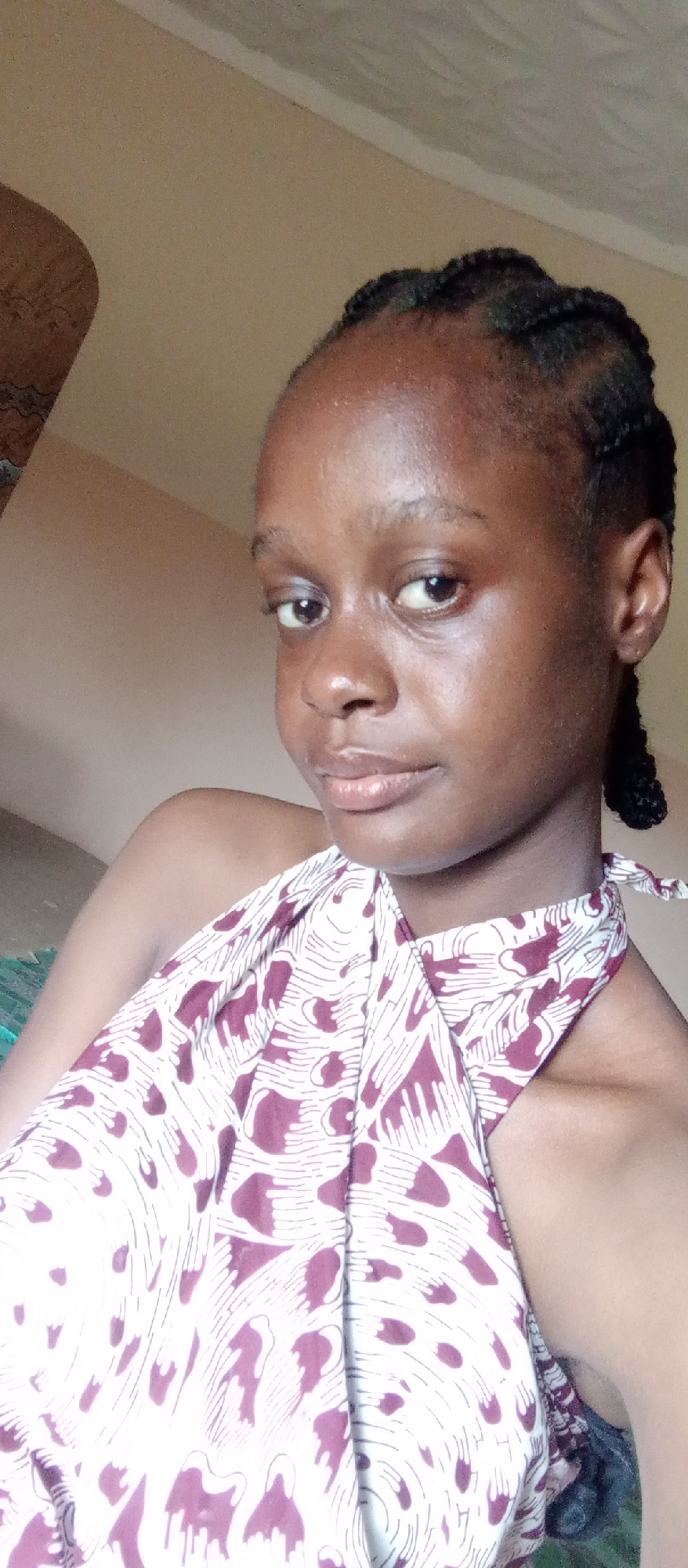 The image is a close-up portrait of a young woman, likely taken indoors in Akwanga, Nigeria. She has dark skin with a visible sheen, especially on her forehead and cheeks. Her dark hair is styled in neat braids, pulled back from her face, with some braids visible around her hairline and behind her ear. She looks directly at the camera with a neutral, calm expression, featuring wide, dark eyes and full lips.
She is wearing a halter-neck top that is primarily white, adorned with a repeating pattern of abstract maroon or dark red shapes, some resembling hands or organic forms, connected by thin white lines. Her right shoulder and upper arm are bare and visible in the foreground.
The background is a plain, light brown or beige wall, with a slight texture visible on the upper parts, possibly near a ceiling line. The lighting is soft and even, suggesting it was taken during the day. There is no visible text or specific location cues beyond the plain indoor setting. The overall mood is direct and composed.