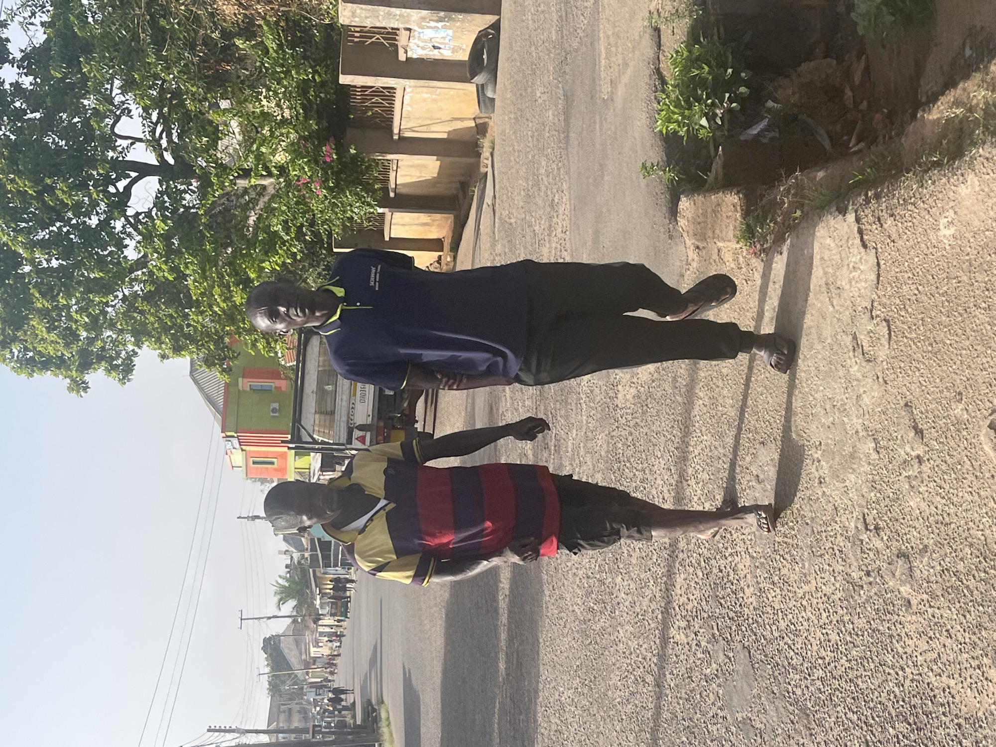 Two African men stand on a concrete sidewalk next to a paved road in Jos, Nigeria. The man in the foreground wears a red and purple striped shirt with gold sleeves and dark shorts. The man behind him is wearing a dark blue polo shirt with dark pants. Buildings and trees line the street in the background. Power lines stretch across the sky.