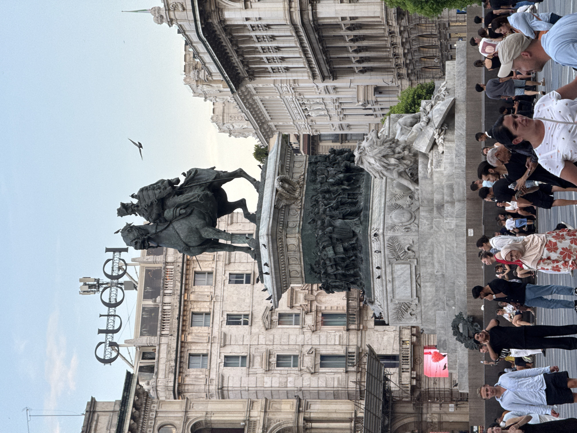 The image depicts a daytime scene in Piazza del Duomo, Milan, Italy. Dominating the center is the equestrian statue of Vittorio Emanuele II, a monumental bronze sculpture atop a large marble pedestal adorned with reliefs and lion figures. In the background, the iconic GUCCI logo is prominently displayed on a building, identifiable by its distinctive font and surrounding architecture featuring numerous windows and balconies. The square is populated with a diverse group of people, some walking, others gathered in conversation, and some observing the surroundings. A few birds are visible in the sky, one in flight near the statue. The overall atmosphere suggests a public space bustling with activity.