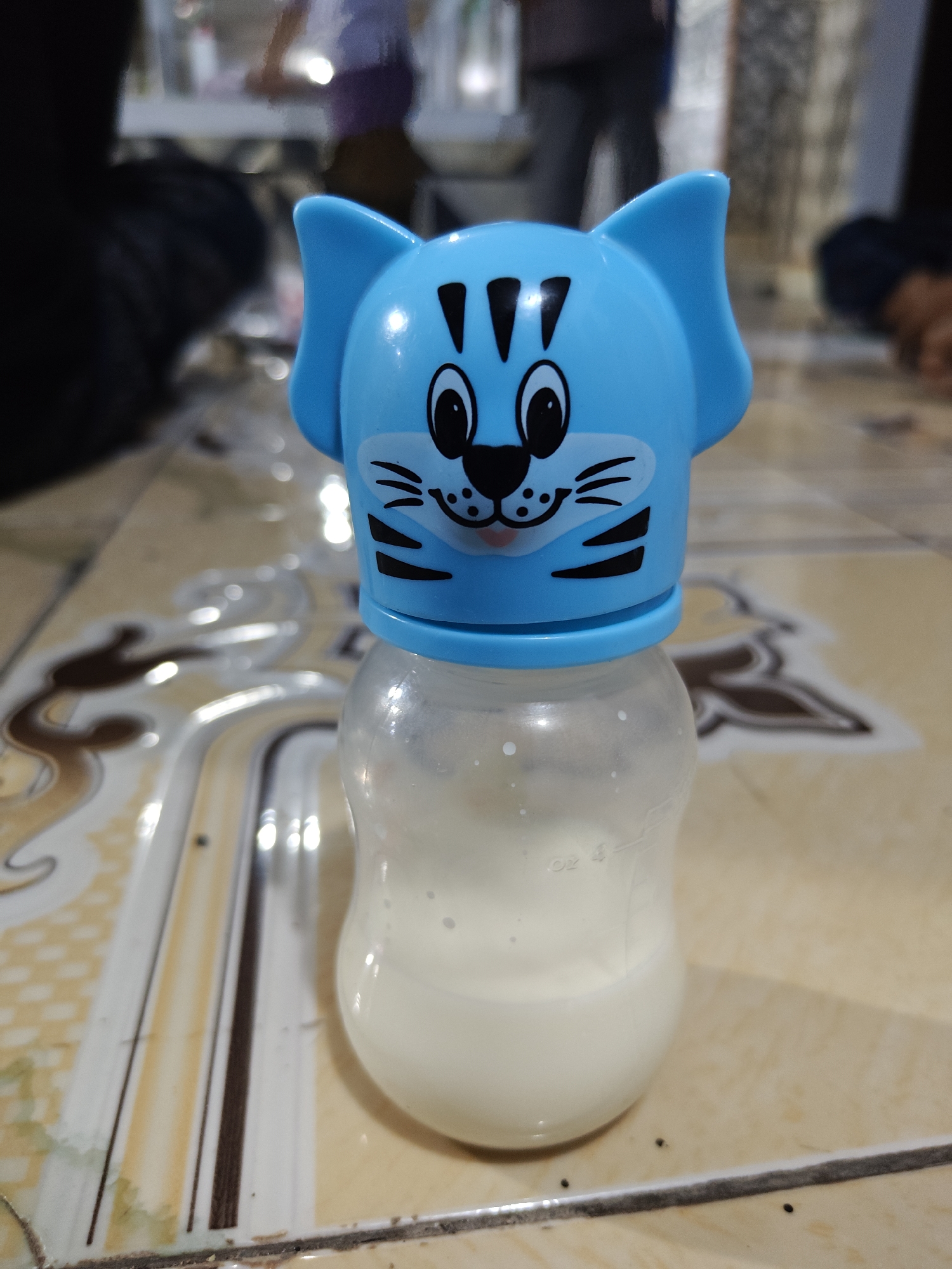 A close-up, eye-level, vertical shot shows a blue cat-themed baby bottle filled with white liquid, possibly milk, sitting on a tiled floor. The bottle's cap is shaped like a cartoon cat face with black markings for eyes, nose, mouth, and whiskers. The bottle has measurement markings on its side. The background is blurred but shows hints of people in a dimly lit indoor setting. The floor tiles are light brown with a pattern of darker brown and yellow lines. The scene appears to be in a home or casual indoor space in Klampokan, Indonesia.