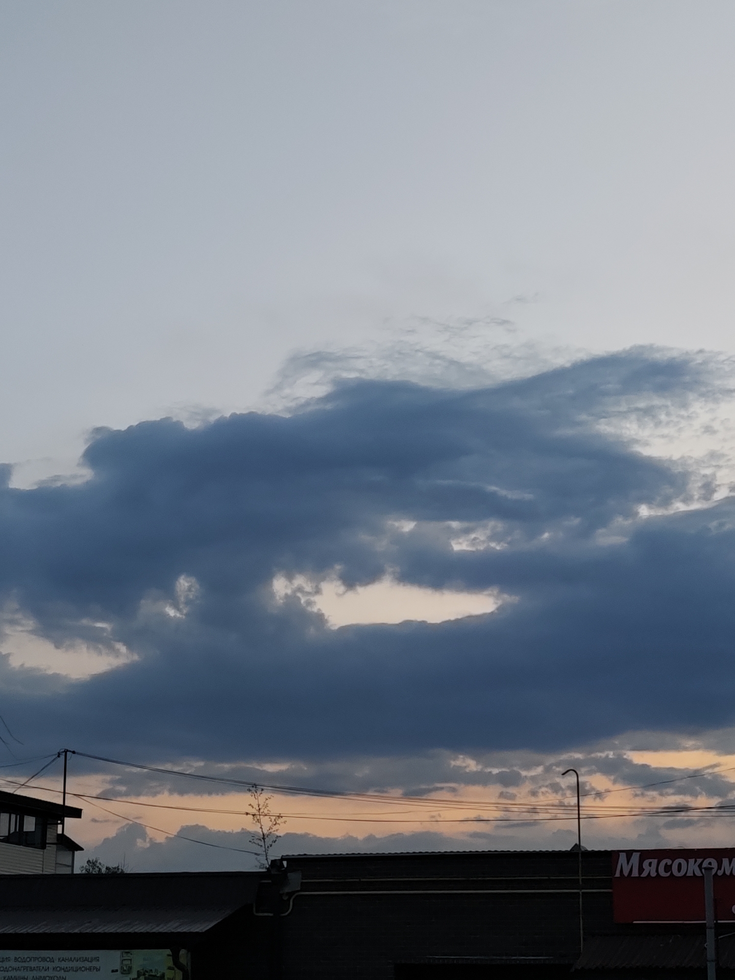The image depicts a cloudy sky over a commercial area, likely in the city of Chaikovsky, Russia, based on the Cyrillic text on the building sign that reads "Мясоком" (Myasokom, likely short for "Meat Combine"). Below the clouds, the skyline includes a building with a red sign for Myasokom and the rooftop of a shorter building with signage in Russian on the left. A utility pole with wires and a curved streetlight are visible against the backdrop of the sky. The sky appears to be at dusk or dawn, with hints of pale orange near the horizon.