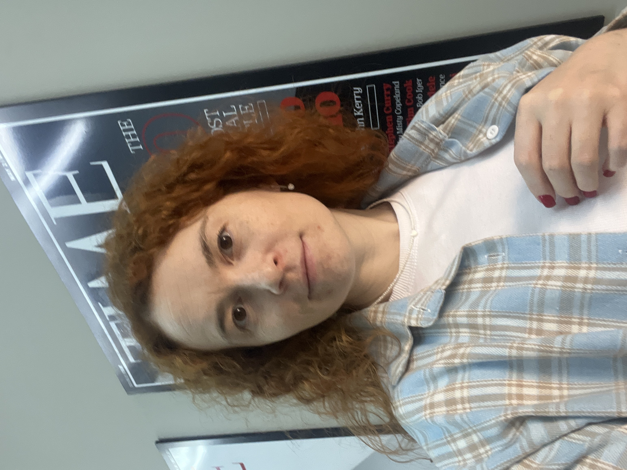 The media file presents a close-up, portrait-oriented image of a person, likely a woman, with reddish-brown, curly hair. She looks directly at the camera with a neutral to slightly contemplative expression, her lips slightly parted. Her skin is fair with visible freckles.
She wears a light-colored, possibly white, inner top beneath a light blue, white, and beige plaid flannel shirt. Her left hand is visible near her chest, displaying red nail polish. A small white stud earring is visible in her right ear.
The background is an indoor setting, likely a public or institutional space such as an office or school in Chaikovsky, Russia. It features a dark-colored poster or display board with prominent white and red text. This poster appears to be a list or article detailing influential figures.
Visible text on the poster includes fragments like "THE," "OST" (likely part of "MOST"), "AL," and "LE." More complete names and phrases are clearly discernible, such as "in Kerry," "Stephen Curry," "Misty Copeland," "Tim Cook," "Bob Iger," and "Adele." The arrangement of the text strongly suggests a list of "The Most Influential" people, possibly akin to a publication's annual feature.