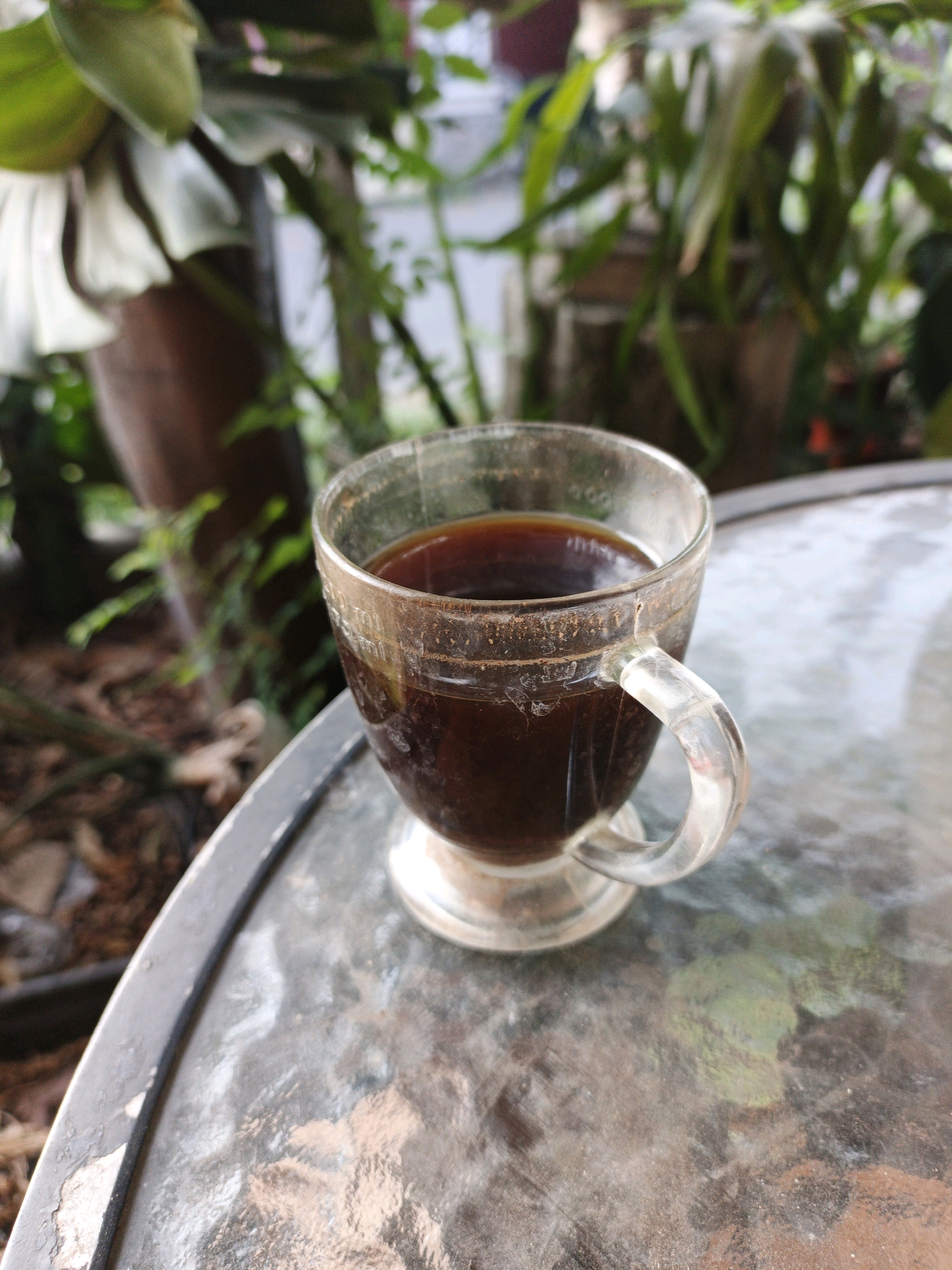 A clear glass cup filled with dark brown liquid, likely coffee, sits on a textured glass table outdoors. The cup has a clear glass handle and is partially submerged in the liquid. The table's surface reflects the surrounding environment, which includes greenery and possibly potted plants. The background is blurred, showing vegetation and possibly some structures. The overall impression is a still-life captured in an outdoor setting, possibly a patio or garden area, located in Ketegan, Indonesia.