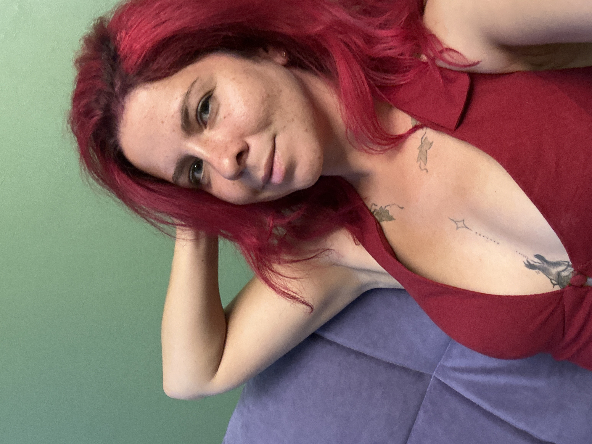 A close-up, eye-level portrait captures a woman with striking, long, wavy red hair and fair skin, adorned with numerous freckles across her face. She has light-colored eyes and a gentle, relaxed expression with a subtle smile, gazing directly at the viewer. Her head is tilted and appears to be resting on her left arm or a soft, textured purple surface, which occupies the lower right portion of the frame. She is wearing a deep red top or dress with a V-neckline. Several small, faded tattoos are visible on her upper chest and shoulder area, including abstract designs and what resembles a small bird or creature near her armpit. The background is a plain, light green wall, suggesting an indoor setting. The lighting is soft and natural, indicating daytime. The image conveys a calm and approachable mood. This photograph was taken in Novosibirsk, Russia.