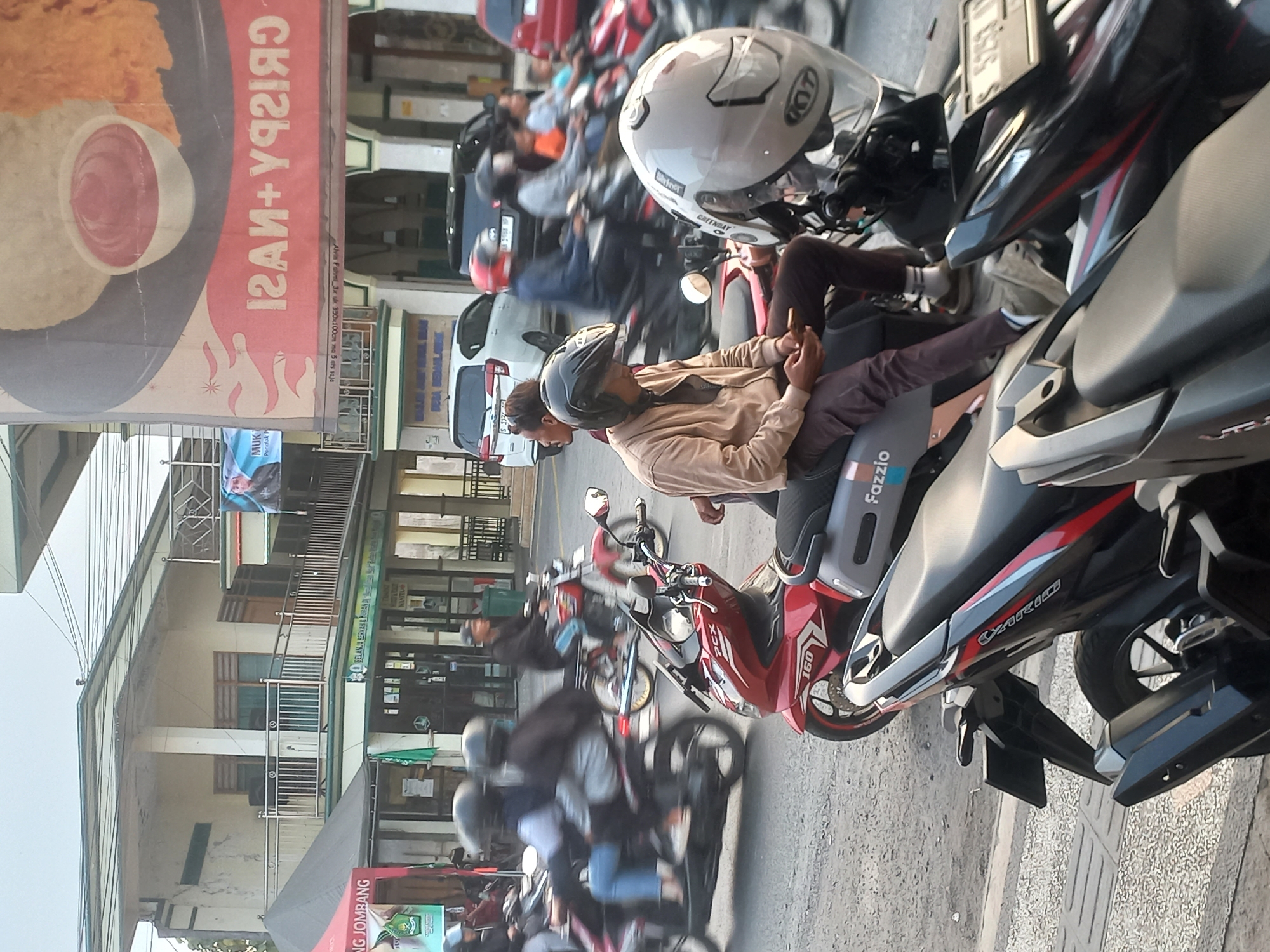 The image depicts a street scene in Bedah Lawak, Indonesia, with several motorcycles and people. In the foreground, a man wearing a tan jacket and dark pants, with a black helmet, is seated on a light grey motorcycle, looking at his phone. Next to him is a red and black motorcycle. Further back, multiple motorcycles are in motion, with riders wearing helmets. Buildings line the street, one with a large banner displaying text and an image of food. Another building has a balcony with railings. Various signs are visible on the buildings, including one in green and white text. The environment appears to be an urban street with moderate traffic.