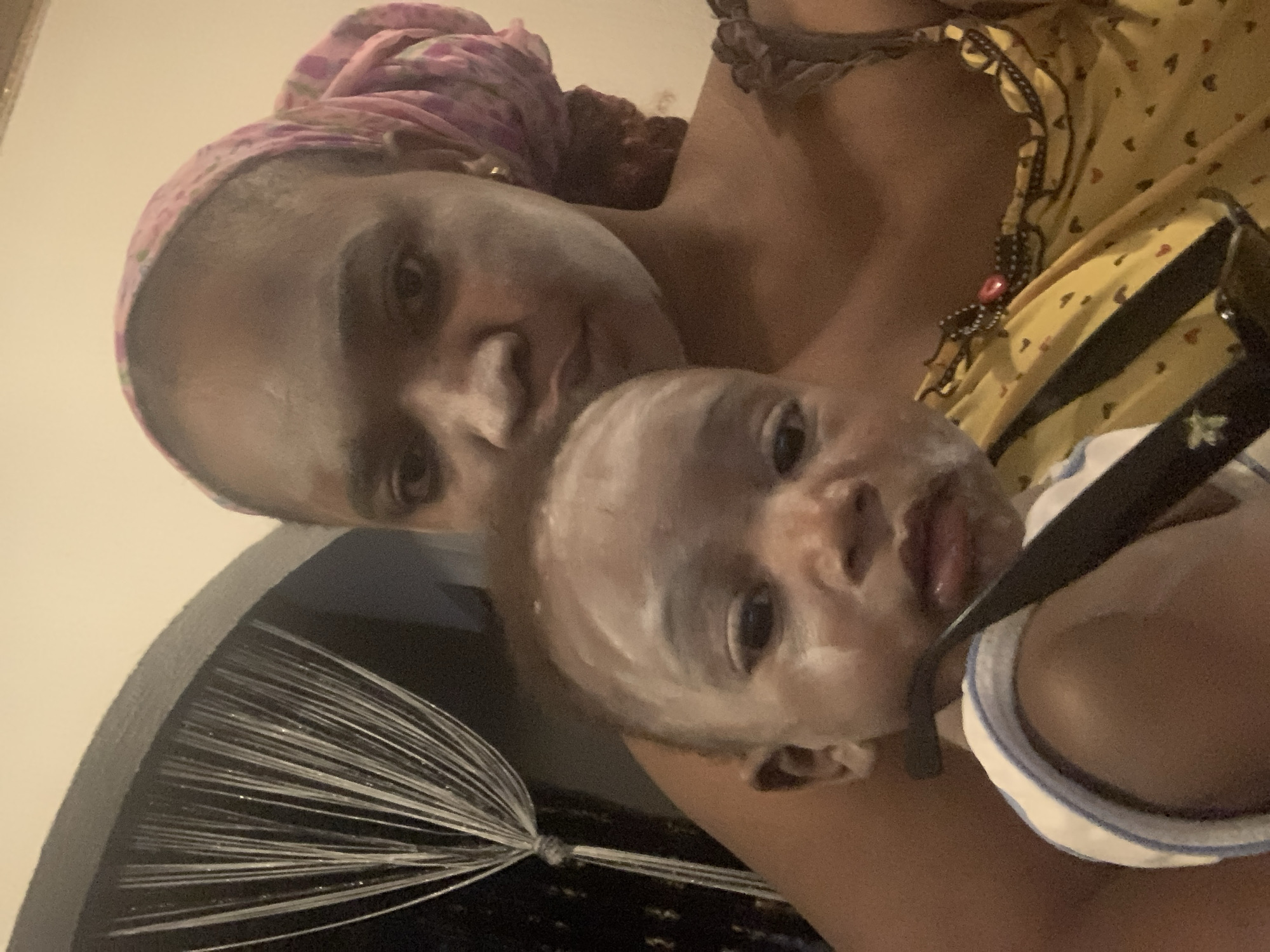 A mother is holding her baby. Both have white cream on their faces. The mother wears a pink headscarf and a yellow top with black and red accents. The baby is wearing a white and blue outfit. The setting is indoors, with a decorative object featuring a dark surface and white radiating lines visible in the background. The city of Barakin Akawo, Nigeria is not discernible from the image.