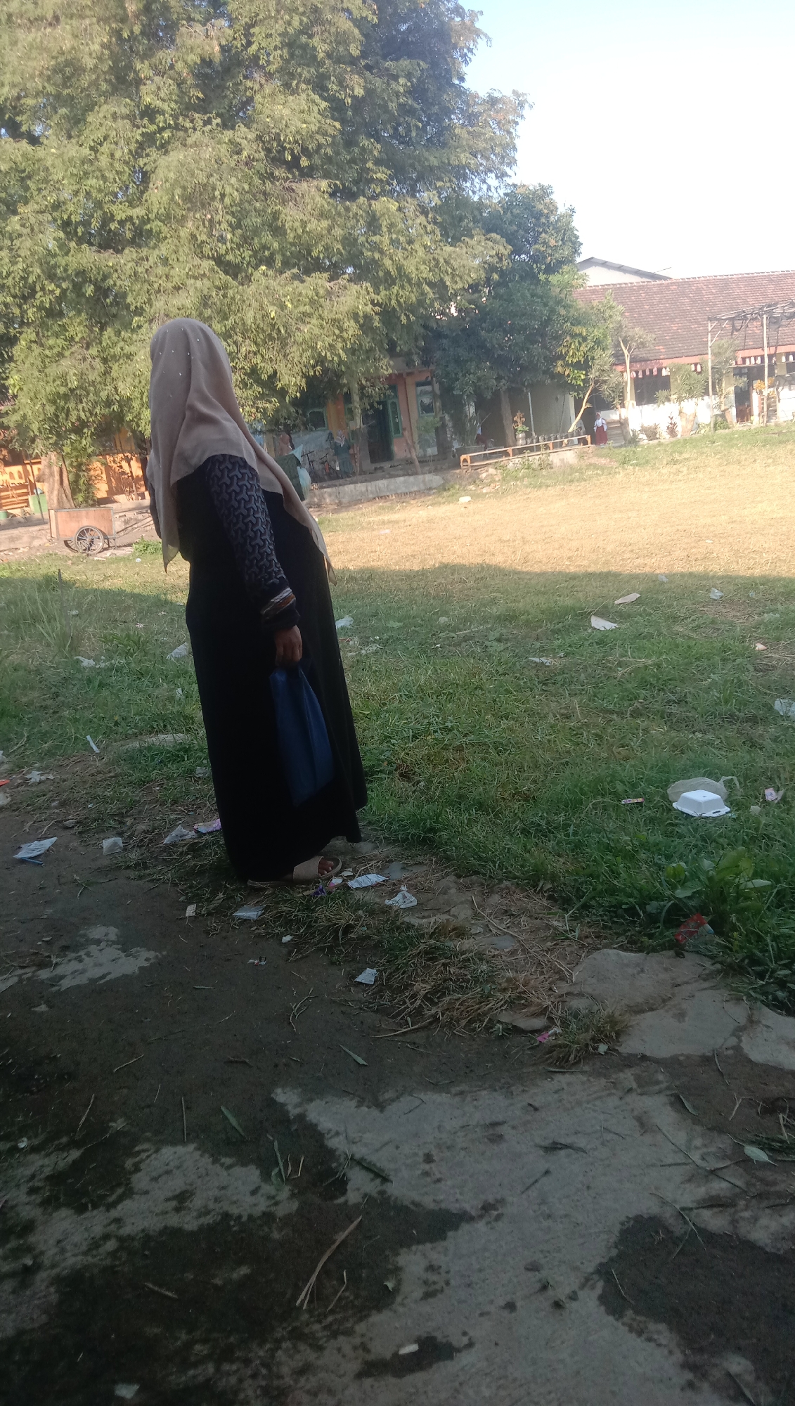 A person wearing a light brown hijab and a black abaya stands facing left in a grassy area. The person is holding a blue bag in their right hand. The ground in front of the person is a mix of dirt and grass, with some scattered litter. In the background, there are trees, a building with a tiled roof, and some other structures. The scene appears to be outdoors, possibly in a park or schoolyard in Karanganyar, Indonesia. The lighting suggests it is daytime, with sunlight illuminating the scene from the right.