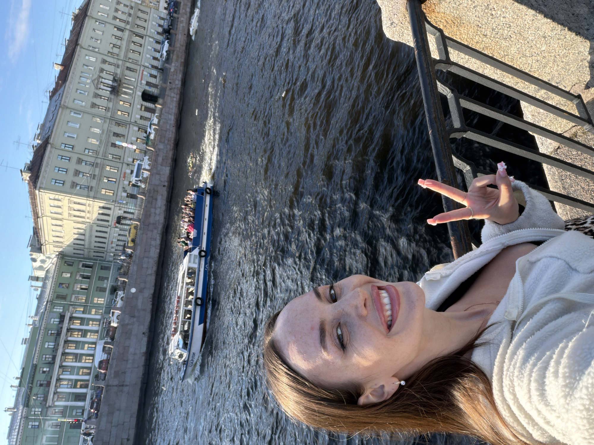 In Saint Petersburg, Russia, a young woman with light brown hair and freckles takes a selfie by a canal. She is wearing a white fleece jacket and a necklace, and she is smiling and making a peace sign with her right hand. Behind her, a tour boat filled with people cruises down the dark water. On the far bank of the canal, multi-story buildings with classical architecture line the waterfront. The sky is bright blue with a few faint clouds.