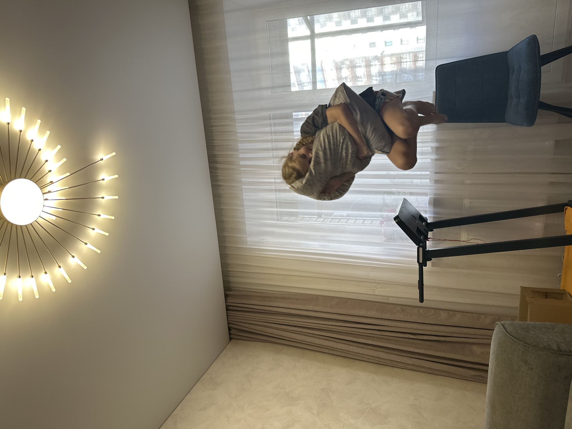 In an indoor setting, a young girl with blonde hair is captured mid-air, holding a pillow. She is wearing a dark t-shirt and patterned shorts, and appears to be jumping. Below her, a dark blue chair and a black treadmill are visible. The room is lit by a unique ceiling fixture with radiating bulbs and natural light coming through sheer white curtains. The walls are textured, one with a light beige stone-like pattern and another covered by beige and brown curtains. The overall scene suggests a playful moment within a residential space. The location of Abakan, Russia, is not visually represented in the provided image and cannot be confirmed.