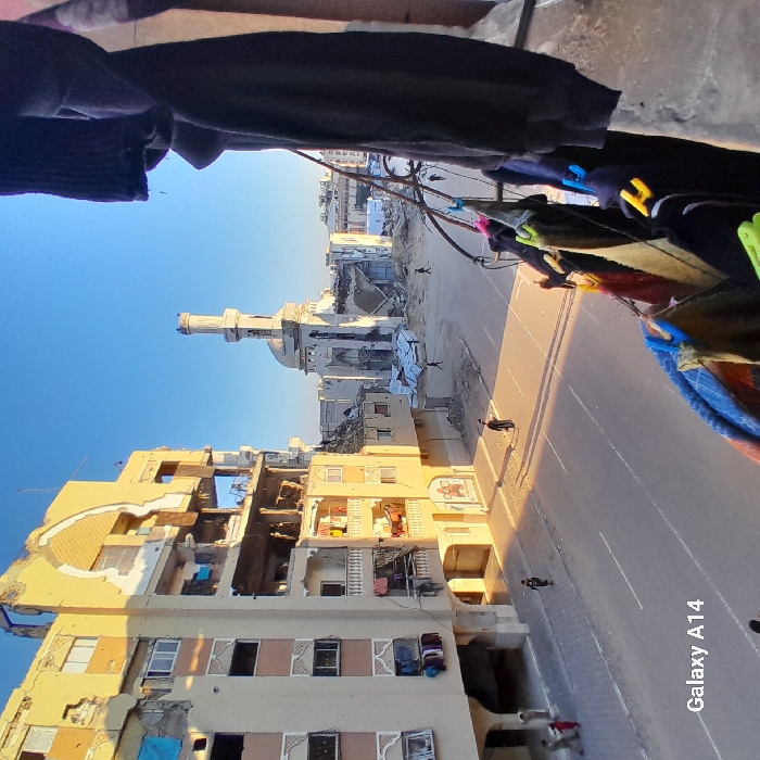 The image presents a wide view of a war-torn urban landscape in Khan Yunis, Palestinian Territory, captured from a slightly elevated vantage point, possibly a balcony. The perspective is rotated 90 degrees clockwise from a typical upright orientation, showing a clear blue sky on the left.
In the foreground, on the right, several pieces of clothing are seen hanging to dry on a line, indicating everyday life continuing amidst the destruction.
The central and background areas are dominated by severely damaged multi-story buildings, many with sections completely destroyed, exposing their interiors. A prominent mosque with a distinct minaret and dome stands further down the main street, also appearing to have suffered significant damage, with visible structural compromises. Debris is scattered around the buildings and on the street.
A wide asphalt road, seemingly a main thoroughfare, stretches through the scene, with long shadows cast across it. A few small figures of individuals are visible walking on the street and near the damaged structures, appearing to go about their routines. The sunlight suggests either early morning or late afternoon, creating strong contrasts and illuminating the golden hues of some surviving building facades. The sky is bright and clear.
The overall scene conveys a profound sense of devastation and the impact of conflict on civilian infrastructure, yet also hints at the resilience of life as people navigate the ruined environment.
The text "Galaxy A14" is visible in the bottom right corner, indicating the camera used.