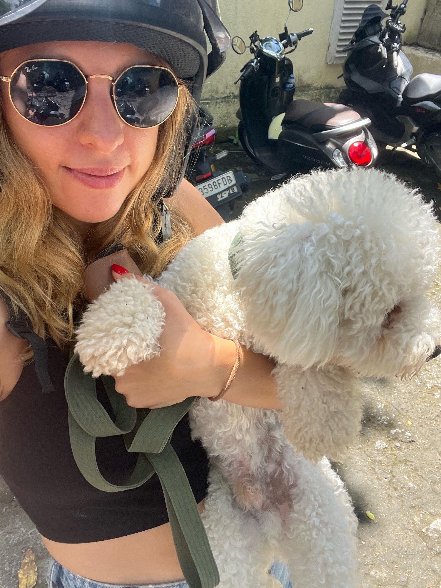 In North Kuta, Indonesia, a woman wearing a black helmet and round, gold-rimmed sunglasses is taking a selfie while holding a white, curly-haired dog. She has blonde hair and is wearing a dark brown tank top. The dog is positioned in her arms, obscuring much of her body. In the background, several scooters are parked, one of which has a license plate that reads "3598FDB". The ground appears to be a paved surface with scattered leaves. The lighting suggests it is daytime.