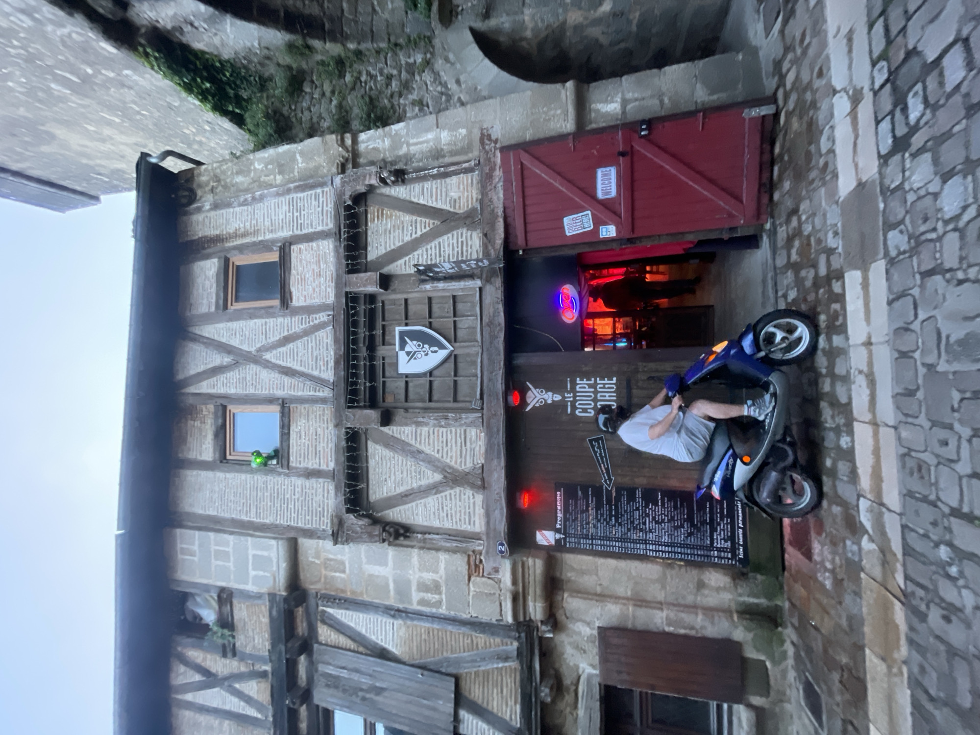 The image shows the facade of a building at 2 Rue de la Vau Saint-Jacques, Parthenay, France. The building is a medieval structure with exposed timber framing and brick infill. A red door provides access to the entrance of "Le Coupe Jarge," a bar or restaurant. A man sits on a blue scooter parked in front of the establishment. A menu board displays the restaurant's offerings. Neon signs and red lights illuminate the entrance. The street is paved with cobblestones.