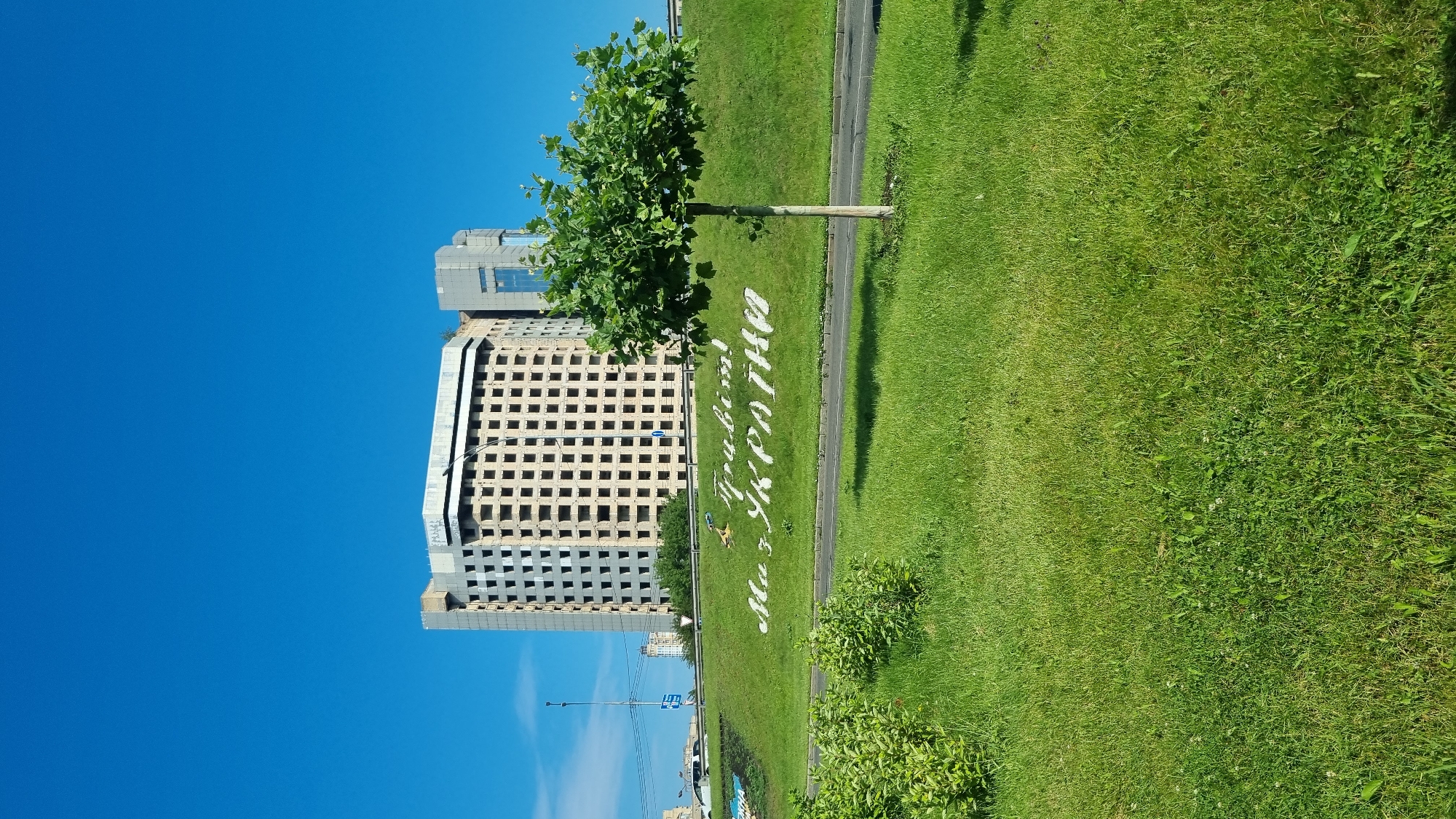 In Kyiv, Ukraine, on a bright, sunny day, a partially constructed or dilapidated building stands against a clear blue sky. The building, an imposing concrete structure with many window frames but no glass, is the focal point of the scene. In front of the building, a lush green hillside slopes down, adorned with white lettering that spells out "Слава Україні!" (Slava Ukraini! - Glory to Ukraine!). The hillside is also dotted with a young, healthy tree on the right side of the frame. A road runs along the base of the hillside, and on the left, there is a patch of dense green bushes. The foreground is dominated by a well-maintained lawn, suggesting a park or public space. The overall impression is one of resilience and national pride, set against the backdrop of ongoing development or reconstruction.