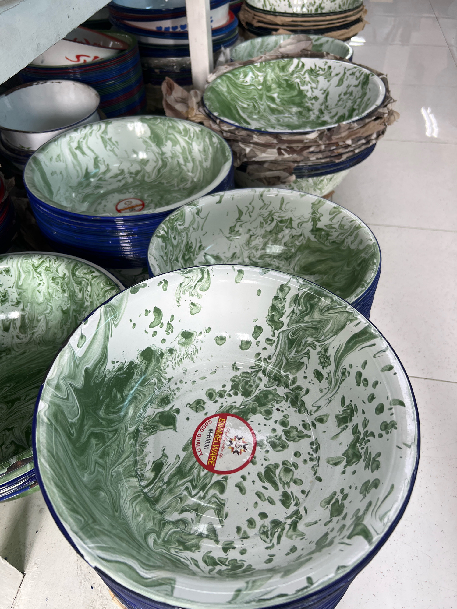 A close-up shot showcases a stack of large, white enamel bowls with a distinctive green marbled pattern, characteristic of Indonesian enamelware, often found in traditional markets or home goods stores. The bowls are rimmed with a dark blue color. In the foreground, the pattern features swirls and splatters of green against a white background. One of the bowls has a red circular sticker with text, including "ENAMELWARE" and "GOOD QUALITY". In the background, more stacked bowls with similar patterns are visible, as well as stacks of colorful striped bowls, predominantly blue and red, suggesting a retail setting. The floor appears to be light-colored tile. The lighting suggests an indoor environment, possibly a shop or market stall in Yogyakarta, Indonesia, given the common availability and style of such goods in that region. There are no people visible in the image.
