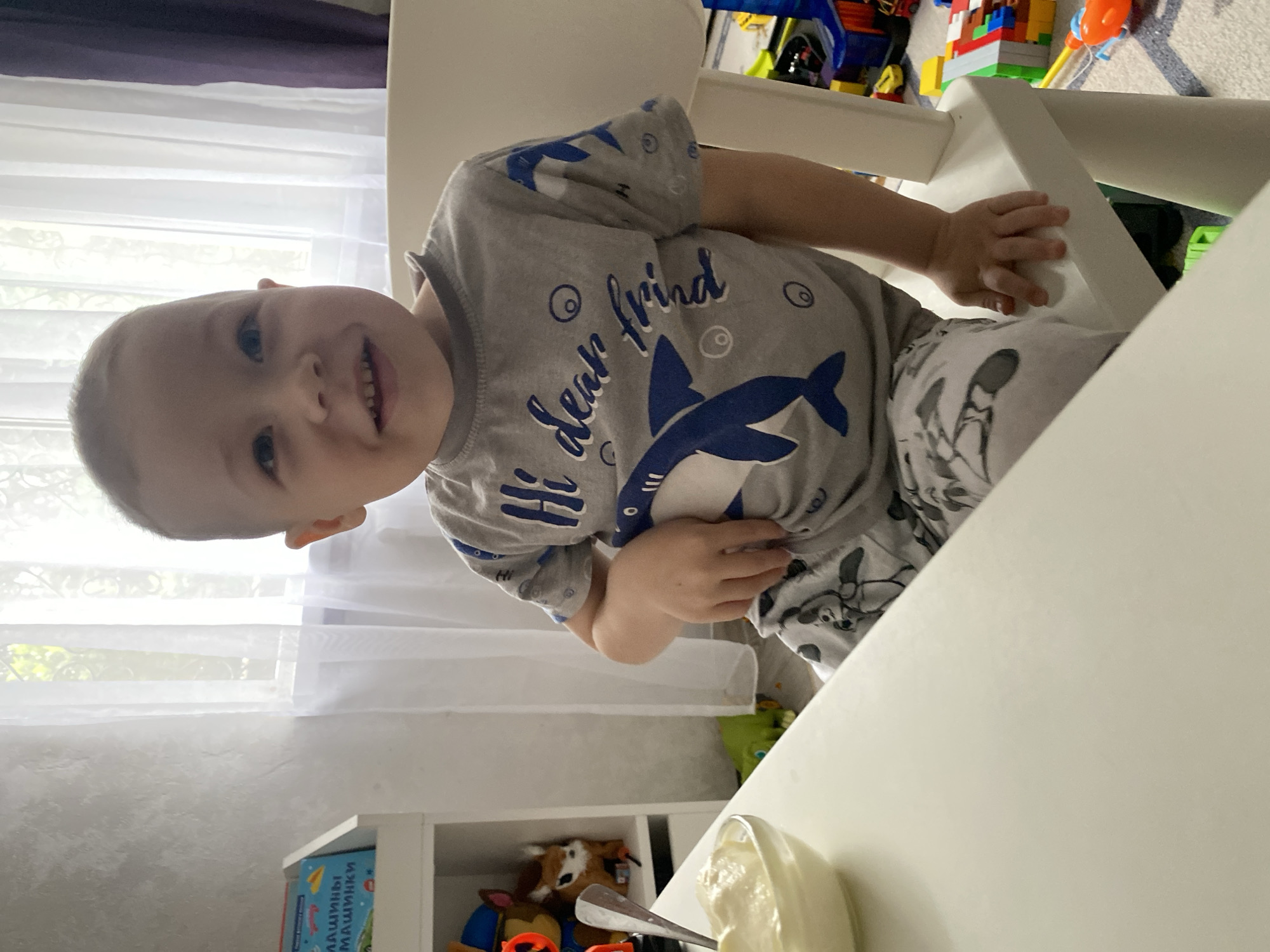 A young child is sitting at a white table, wearing a gray shirt with a shark design and gray pants with a pattern of teddy bear faces. The child is smiling at the camera. A bowl of what appears to be yogurt and a spoon are on the table. Behind the child, there is a window covered with white sheer curtains. To the left of the window, a purple curtain is visible. Below the table, there is a white shelf with children's books and toys. A collection of various toys are visible in the background, with some piled on a rug. The child is sitting in a white high chair. The location is not discernible from the image.