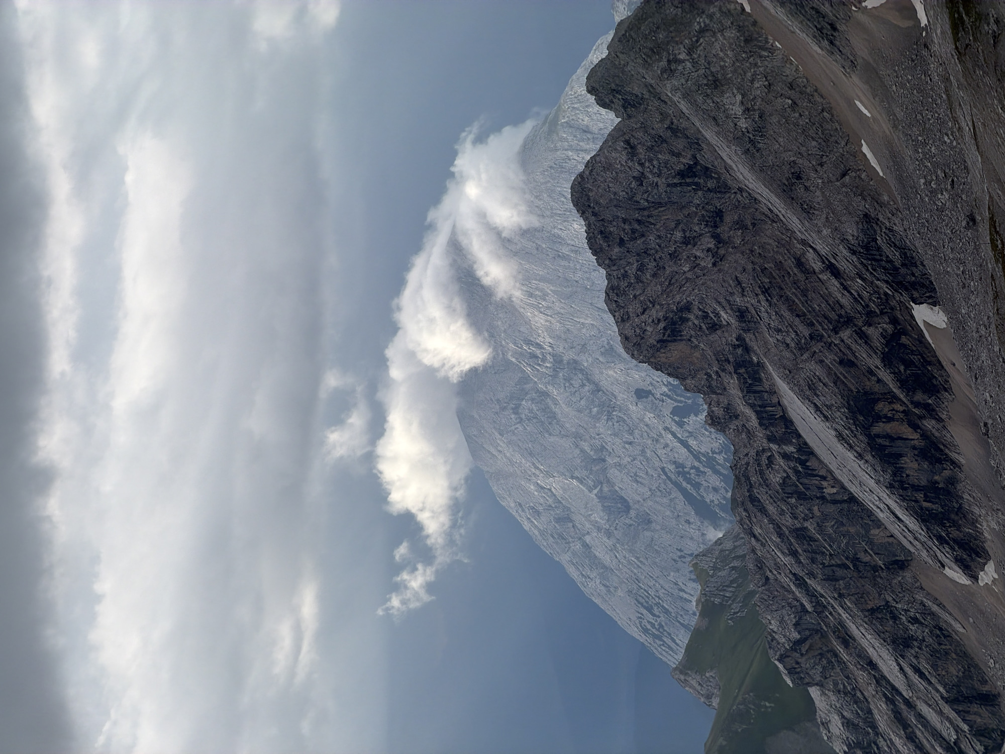 The image depicts a dramatic mountain landscape under a cloudy sky, likely in the vicinity of Garmisch-Partenkirchen, Germany, a region known for its alpine scenery. The foreground features rugged, dark-colored rock formations with steep inclines, suggesting a high-altitude environment. Sparse patches of snow are visible on the rocky slopes.
In the mid-ground, a prominent, lighter-colored mountain peak rises, partially obscured by wisps of white cloud that trail across its summit. This peak appears to be composed of weathered rock, with visible striations and textures.
The background is dominated by a vast, overcast sky filled with layers of gray and white clouds. The lighting is diffused, creating a somewhat moody and atmospheric scene. The overall impression is one of natural grandeur and the raw power of the alpine environment. There are no visible people or human-made structures in this image, focusing solely on the natural elements of the mountainous terrain and sky.