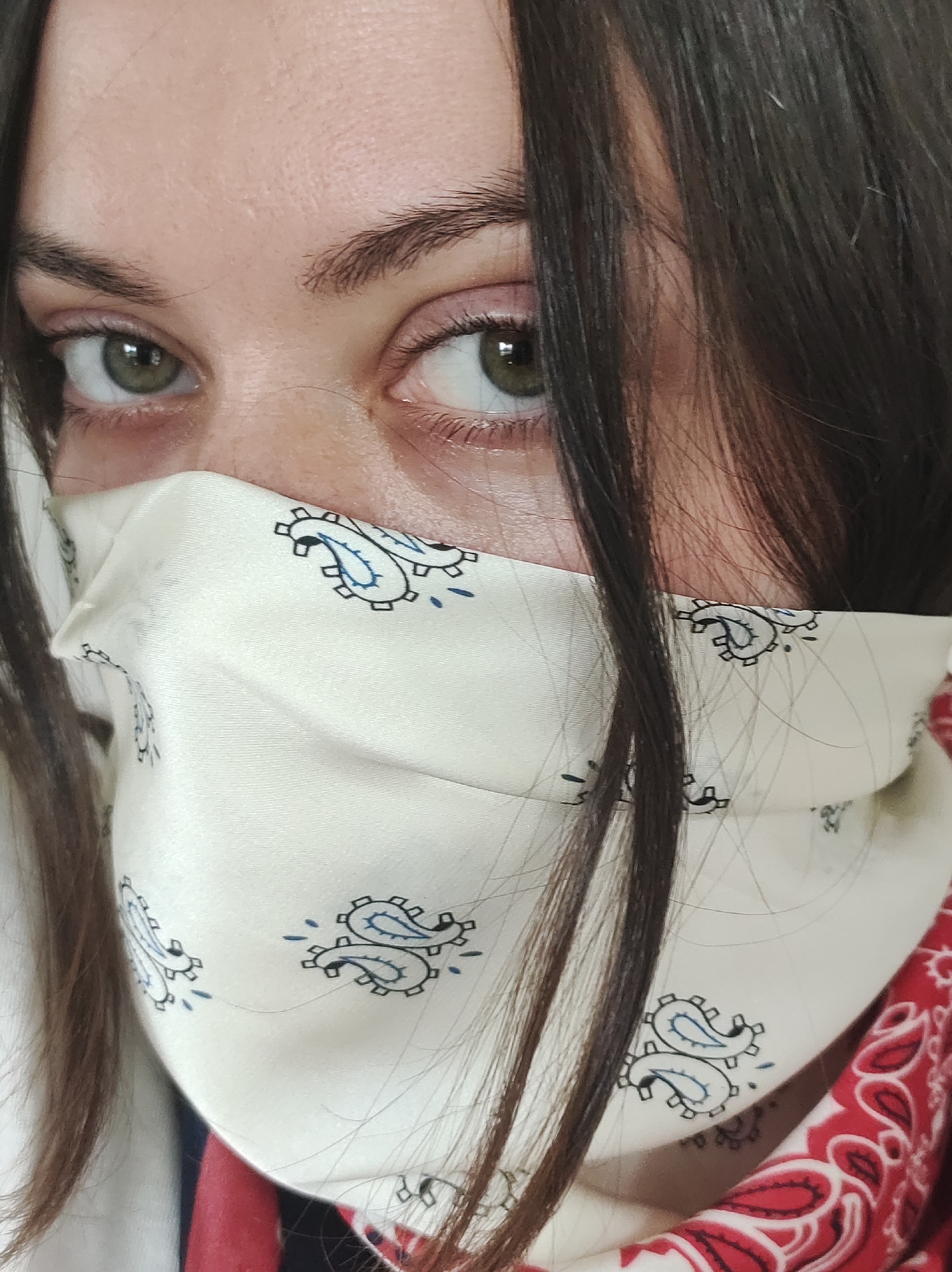 A person's face is partially visible, with the focus on their eyes and the top of a bandana-style face mask. The person has brown hair, some strands of which partially obscure their face. Their eyes are green or hazel. The face mask is a light color with a paisley pattern in blue and black. A red and white patterned fabric is visible behind the light-colored mask, suggesting the layering of another bandana. The image appears to be a close-up selfie. The location cannot be determined to be in Фоки, Russia from the image.