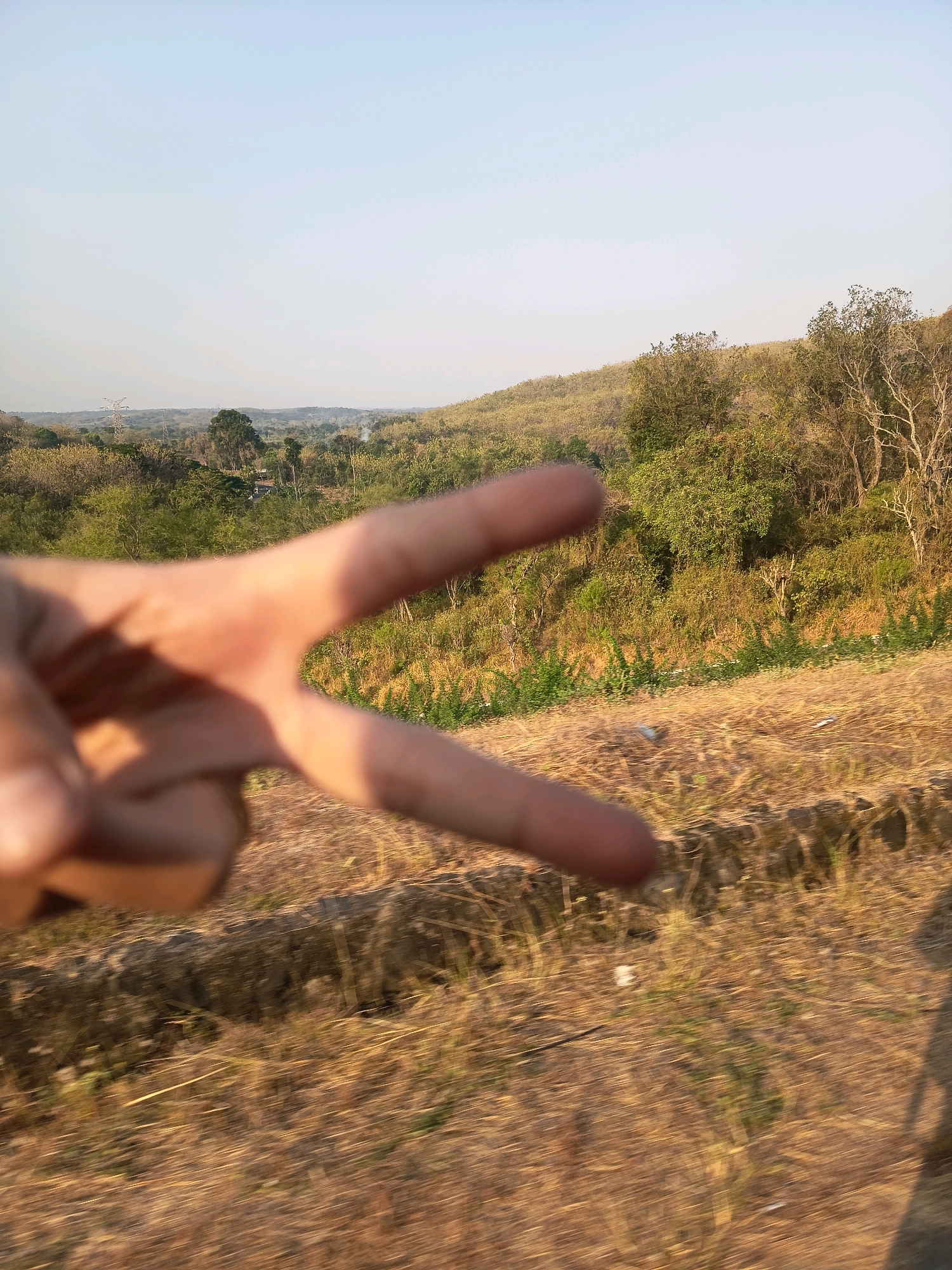 An out-of-focus hand forms a peace sign in the foreground, obscuring a landscape of rolling hills and dry vegetation. The background shows a hazy sky and a distant power line tower, suggesting an open, possibly rural area in or near Kalangbancar, Indonesia. The dry grasses and sparse trees indicate a warm or dry climate.