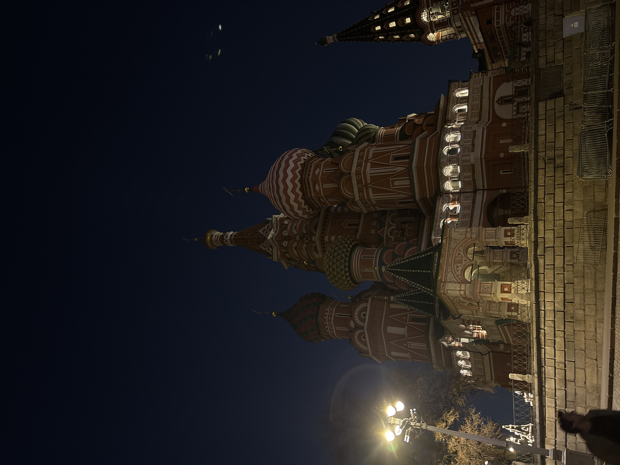 The image shows St. Basil's Cathedral in Moscow, Russia, at night. The cathedral is brightly lit, highlighting its colorful domes and intricate architectural details. The sky is dark blue. A portion of the Kremlin wall and a street lamp are also visible in the foreground. The image is taken from a low angle, looking upwards towards the cathedral.