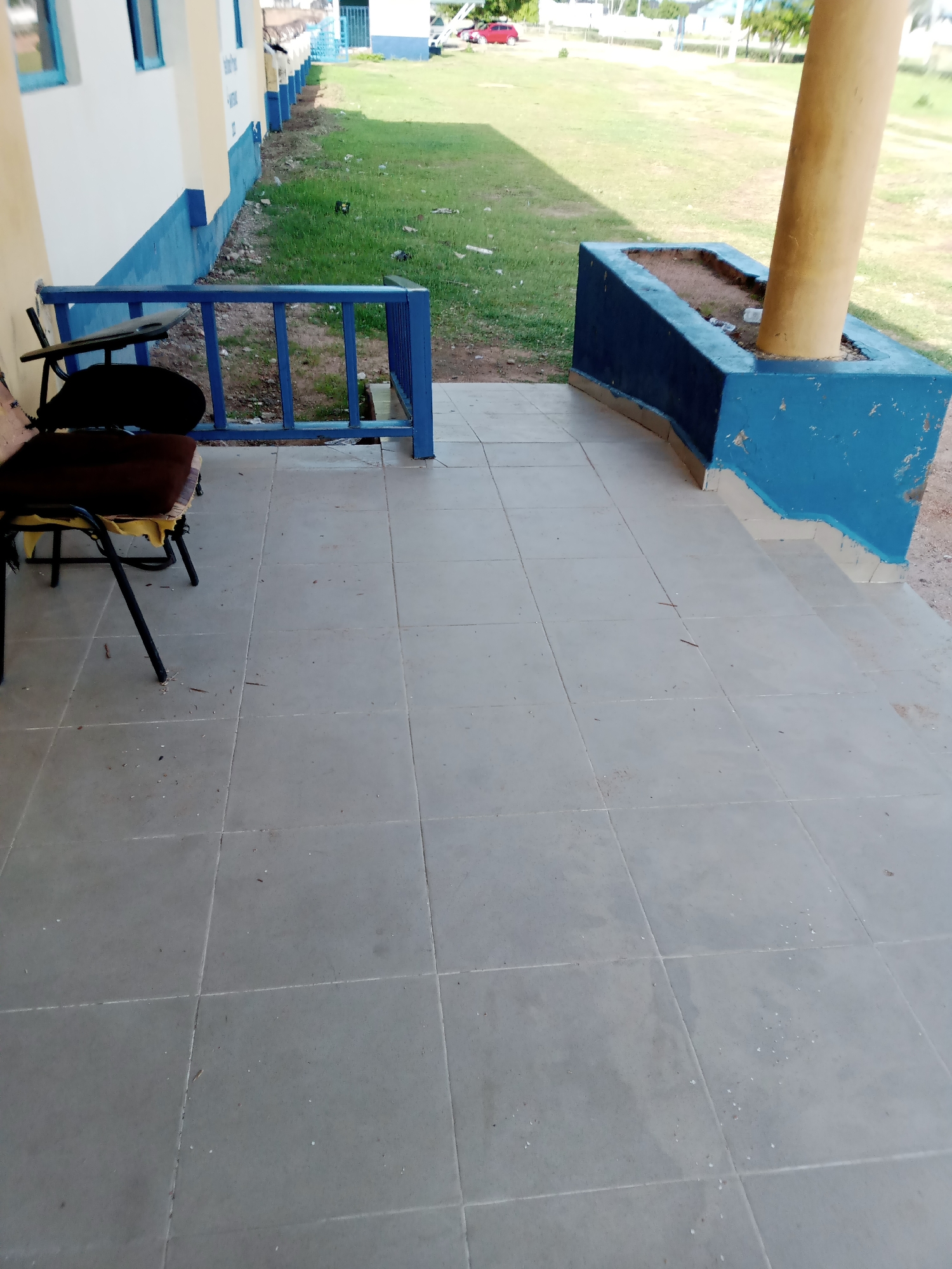 The image depicts an outdoor scene, likely in Jalingo, Nigeria, featuring a tiled patio area in front of a building. A blue metal railing with vertical bars separates the patio from a grassy area and a building. To the left, a dark-colored chair with visible wear and tear sits on the tiled surface. In the background, a large, beige, cylindrical pillar rises from a blue, concrete planter box filled with soil and debris. The building itself has white walls with blue trim and blue-framed windows. The ground is covered in light gray square tiles. The overall impression is of an outdoor space adjacent to a building, possibly a school or public facility, showing signs of outdoor wear and tear.