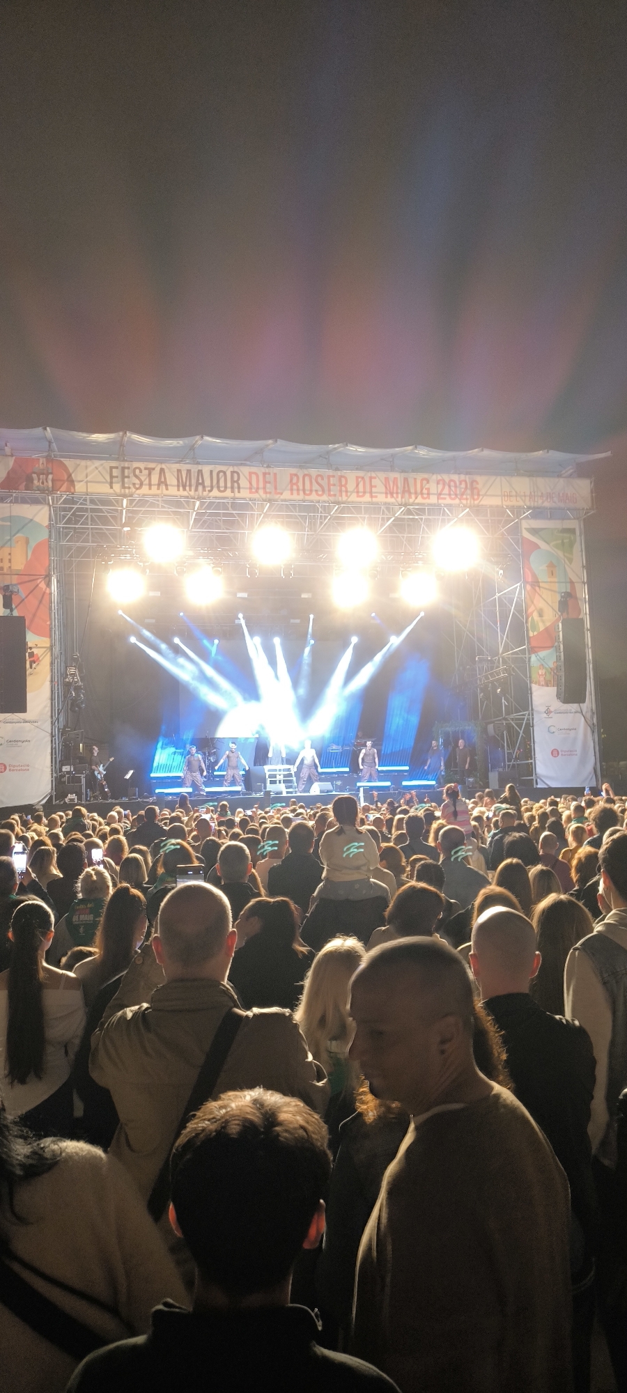 The image shows a large crowd of people gathered at night to watch a live musical performance on a stage with bright lights. Above the stage, there is a banner that reads "Festa Major del Roser de Maig 2026." The scene is in Cerdanyola del Vallès, Spain, as indicated by the signage and event name. Several figures are on the stage. The people in the crowd are facing the stage, watching the performance. The environment appears to be an outdoor venue.