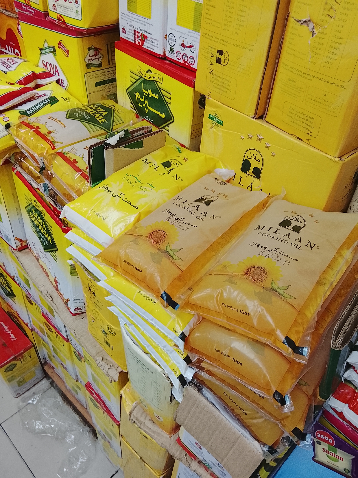 The image shows a stack of cooking oil and vegetable ghee products in a retail setting. The dominant brand is "Milaan" cooking oil, packaged in yellow bags with sunflower imagery and text in both English and Urdu. Several bags of "Soya Supreme" cooking oil and ghee are also visible. The location may be inferred from the presence of the Urdu language on product labels and the potential association with the Gulgasht Colony in Pakistan due to the presence of regional-specific brands. The products are stacked and shelved, suggesting a retail environment like a grocery store or supermarket. There are no people visible in the frame. The environment is a typical retail setting with products stacked for display. The image appears to be a stock photo of common cooking products.