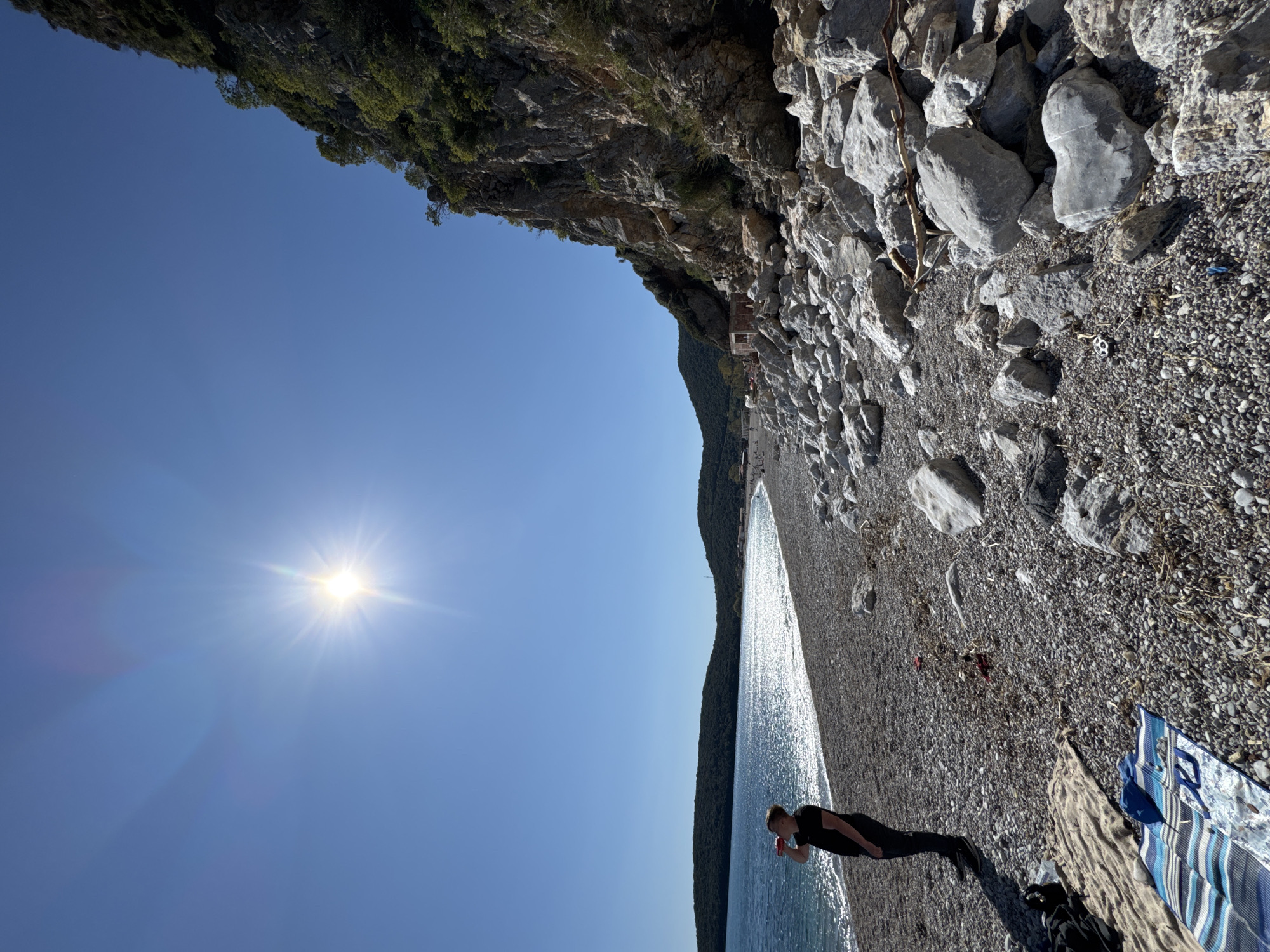 A wide-angle shot oriented sideways shows a sunny day at a stony beach in Prijevor, Montenegro. The water is sparkling, and a figure wearing black is standing at the edge of the water, looking down. A blanket and backpack are on the shore nearby. Above the beach are boulders and a cliff with vegetation. The sun is high in the clear blue sky.