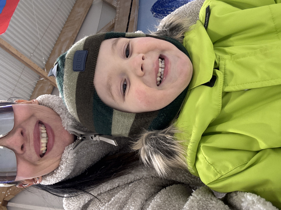 A close-up, chest-level portrait depicts an adult female and a young child, both smiling directly at the viewer, in what appears to be an indoor or covered winter sports facility in Chaikovsky, Russia.
The adult female, positioned in the upper frame, exhibits long dark hair, multiple ear piercings, and a wide smile revealing teeth. She wears large reflective shield-style sunglasses, a light-colored (possibly off-white or cream) fleece jacket with a prominent silver zipper pull, and a light-colored headband or beanie partially visible above her forehead.
The young child, positioned in the foreground, has fair skin with rosy cheeks and a broad smile displaying teeth. The child is dressed in a dark green, khaki, and brown striped winter hat featuring ear flaps and a dark blue "RUDDY" patch on the front, with a small, light-colored object clipped to the top of the hat. A bright neon green winter jacket, complete with a brown and grey faux fur-lined hood, covers the child's torso.
The background shows a sloped, light-colored ceiling with exposed wooden support beams. To the upper right, a partial view of a red and blue banner or flag is visible, alongside a blue and white structure resembling artificial snow or ice. This environment suggests a setting such as an ice rink, ski lodge, or other winter recreational venue within Chaikovsky.