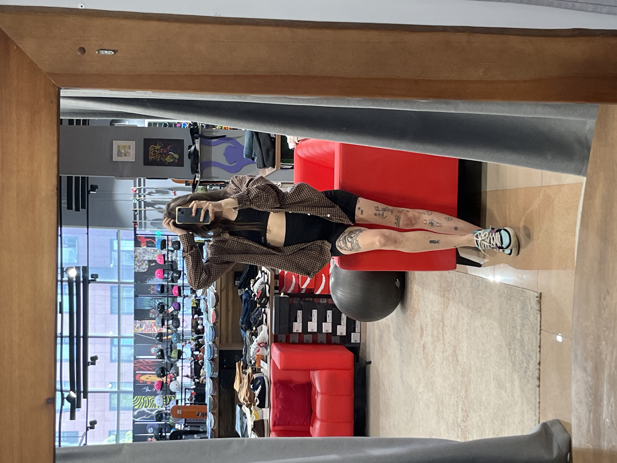 A person wearing a brown plaid jacket, black top, and black shorts, is sitting on a red cushioned chair in what appears to be a dressing room or a retail setting. The person is holding a phone, presumably taking a photo of themself in the mirror. Tattoos are visible on their legs. The room contains a gray exercise ball, shelves stocked with items in boxes, and a wall filled with assorted headwear. The reflected view through the window reveals a cityscape. The general context of the items sold, and architecture of the building in the background suggest that the picture was taken in Krasnoyarsk, Russia.