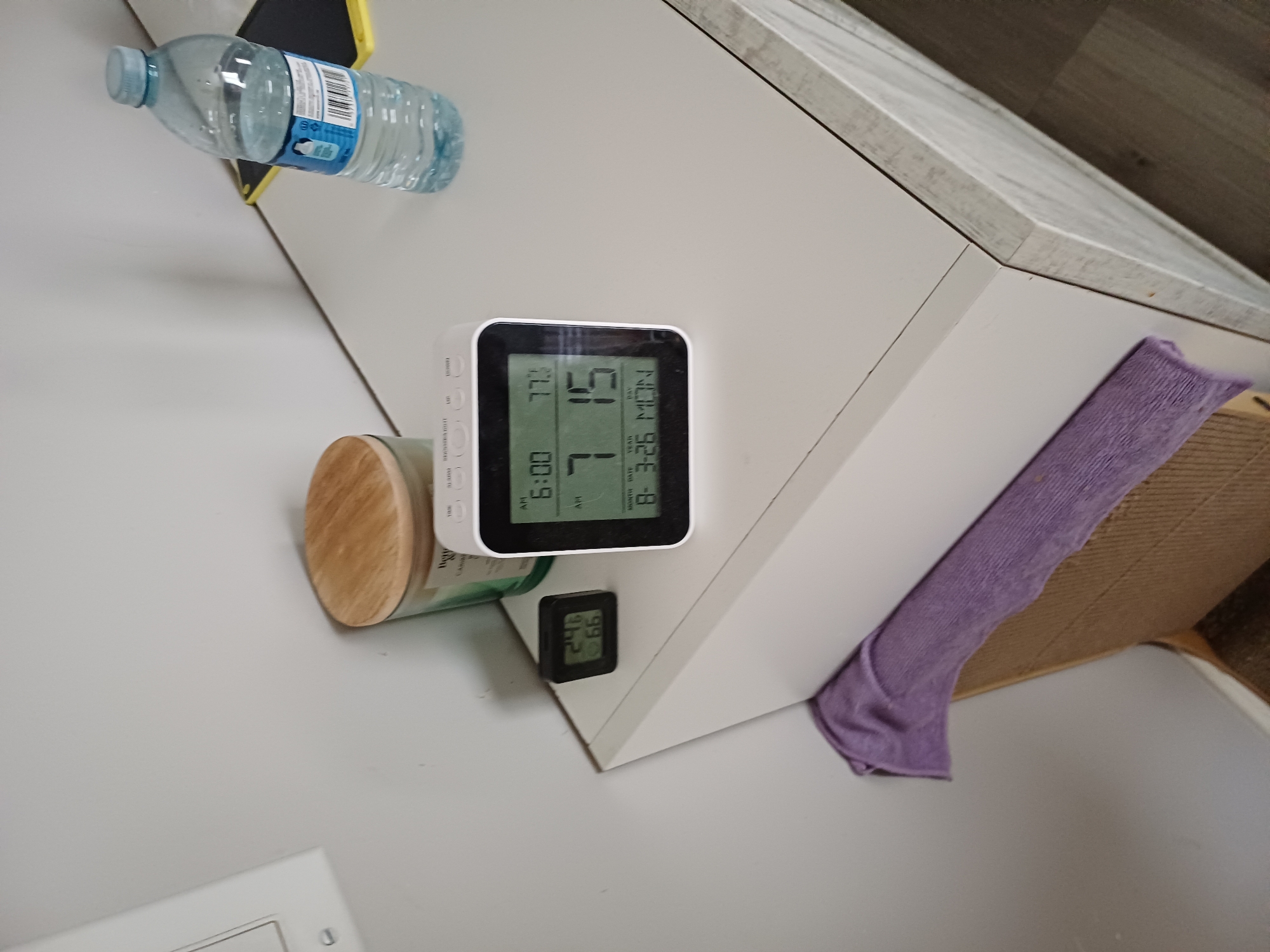 In Morden, Canada, a white nightstand displays various objects. A white digital clock with a black frame shows the time as 6:00 AM, the temperature as 77.3 degrees Fahrenheit, and the date as March 26th, Monday. Next to it, a smaller black digital thermometer reads 24 degrees Celsius and 66% humidity. To the left, a glass jar candle with a wooden lid sits on the nightstand. A clear plastic water bottle with a blue cap and a blue label is positioned on the right side of the nightstand. A folded purple cloth is draped over the edge of the nightstand. The background consists of a plain white wall.