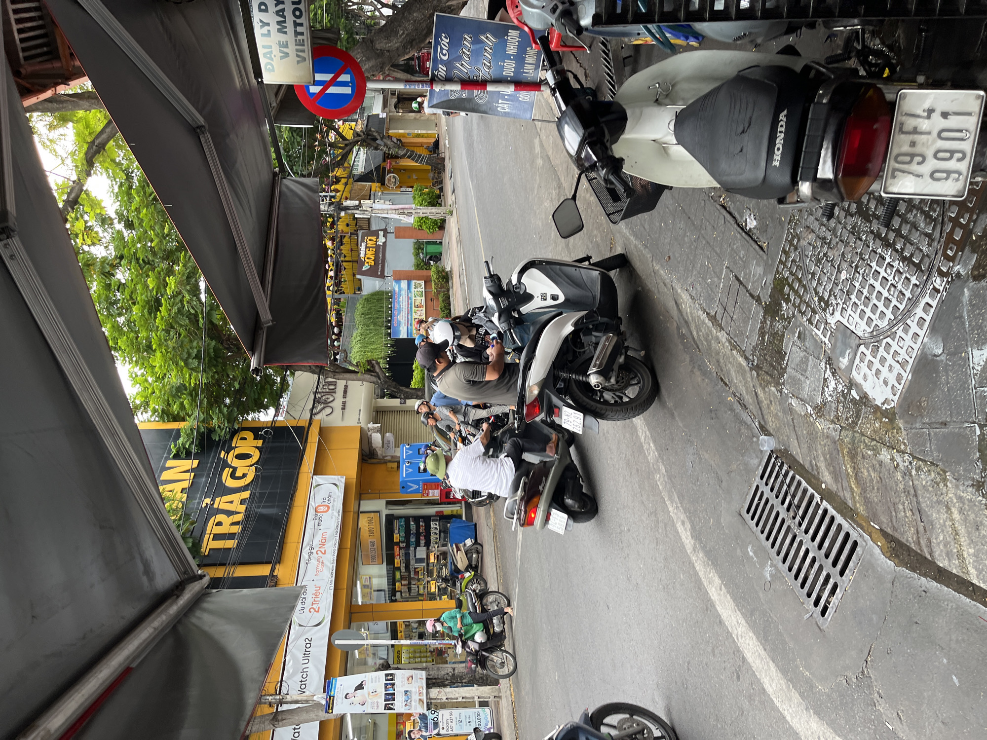 This image captures a street scene in Nha Trang, Vietnam, likely in the daytime given the bright lighting and shadows. The foreground features a section of wet pavement with a metal drain grate and a white and black Honda scooter parked on its side. The scooter's license plate reads "79-FL 9901". Further back, several people on motorcycles are visible. On the left, a person in a green shirt and white helmet rides a dark-colored motorcycle. In the center, two men are on a white scooter, one wearing a helmet and the other a cap. To the right, a man in a light-colored shirt and a green helmet is on a scooter. The background shows buildings with awnings, including one with a large yellow sign that says "AN TRÁ GÓP". Another sign above a storefront reads "DAI LY LI VE MAY VIETTOU". Trees and overhead wires are also present. The overall impression is a busy urban street with common modes of transportation.