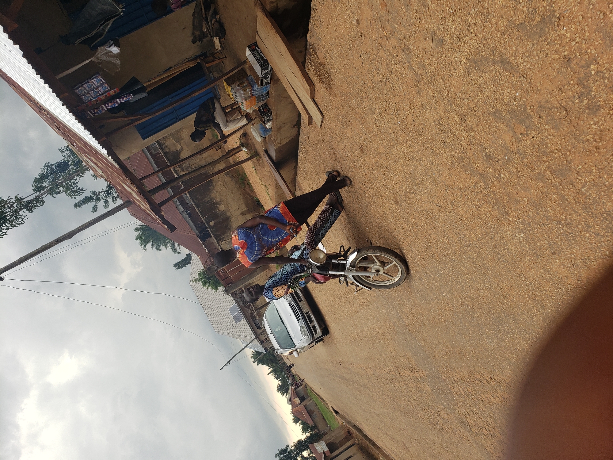The image depicts a street scene in Kachia, Nigeria, under a cloudy sky. In the foreground, two individuals stand beside a motorcycle. One person, wearing a blue and orange patterned sleeveless top and black leggings, leans against the motorcycle. The other person, in a similar blue and orange patterned garment, stands behind the motorcycle, looking towards the left. A silver sedan is parked on the dirt road to the left, with buildings lining the street. To the right, a structure with a corrugated metal roof appears to be a shop or market stall, with goods displayed on shelves and a person seated inside. The road is made of packed dirt and gravel. Trees and vegetation are visible in the background, along with utility poles and wires.