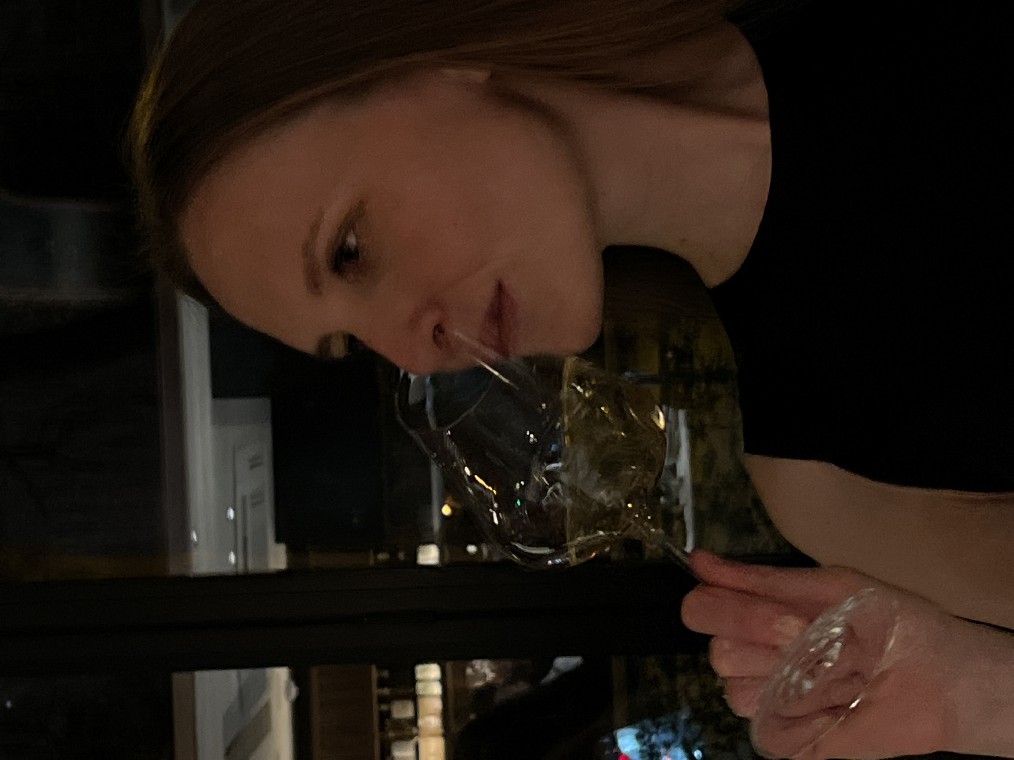 This close-up image, oriented with a slight tilt, features a young woman smelling wine from a glass. She holds a wine glass containing a pale, golden liquid, likely white wine or champagne, with her right hand. Her head is tilted towards the glass, and her nose is close to the rim as she appears to be intently inhaling the aroma. Her expression is focused and calm.
The woman has brown hair pulled back from her face and is wearing a dark, possibly black, top. The setting is indoors, characterized by dim lighting, suggesting an evening or nighttime scene. The background is largely dark and out of focus, but hints of reflections on the glass and blurry lights in the distance suggest an urban environment or an interior with windows overlooking a city or another well-lit area. Some indistinct architectural shapes or shelves are vaguely visible in the background. The lighting highlights the contours of her face and the transparent glass. There is no visible text in the image.
