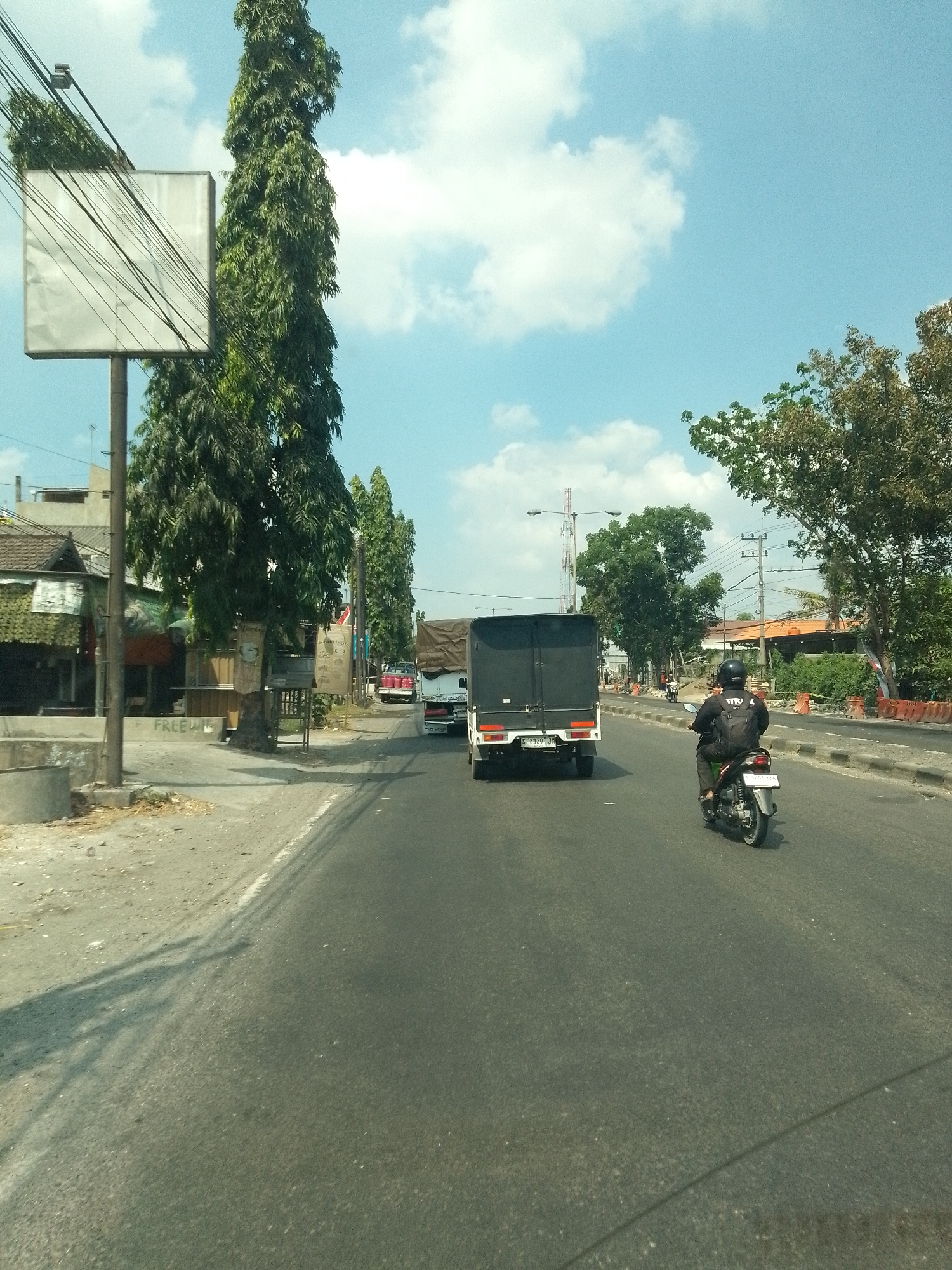 The image depicts a street scene in Plaosan, Indonesia, under a bright blue sky with scattered white clouds. A tall, slender billboard stands on the left side of the road, obscured by numerous electrical wires. Adjacent to it are several tall, leafy trees. The road is paved and appears to be a two-lane street with traffic.
In the foreground and midground, several vehicles are visible. A motorcycle with a rider wearing a black helmet and backpack is traveling away from the viewer on the right lane. In front of the motorcycle, a white truck with a black tarp-covered cargo area is also moving away. To the left of the truck, another truck, possibly a pickup, with a red cabin is seen. On the left side of the road, there are some small buildings and shops with awnings and signage, including what appears to be a sign that reads "FREEW".
The environment suggests a partly urban and partly suburban setting. There are trees lining the road, and some construction barriers with orange cones are present on the right side of the road in the distance. The overall impression is of a typical day on a busy street in Indonesia.