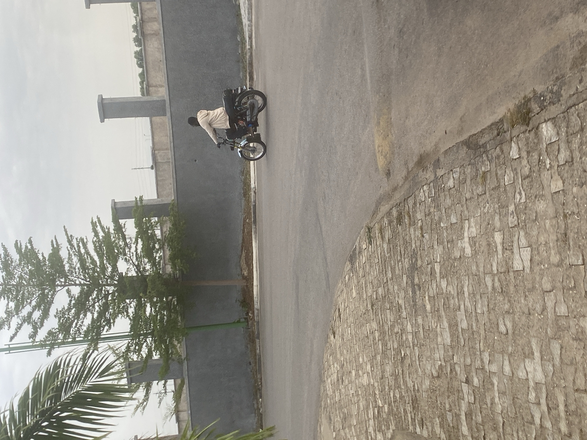 A person wearing a light-colored jacket is riding a motorcycle on a paved road. The road is bordered by a gray wall on one side and a stone embankment on the other. A tree with green foliage is visible behind the wall. The image was taken in Abuja, Nigeria.