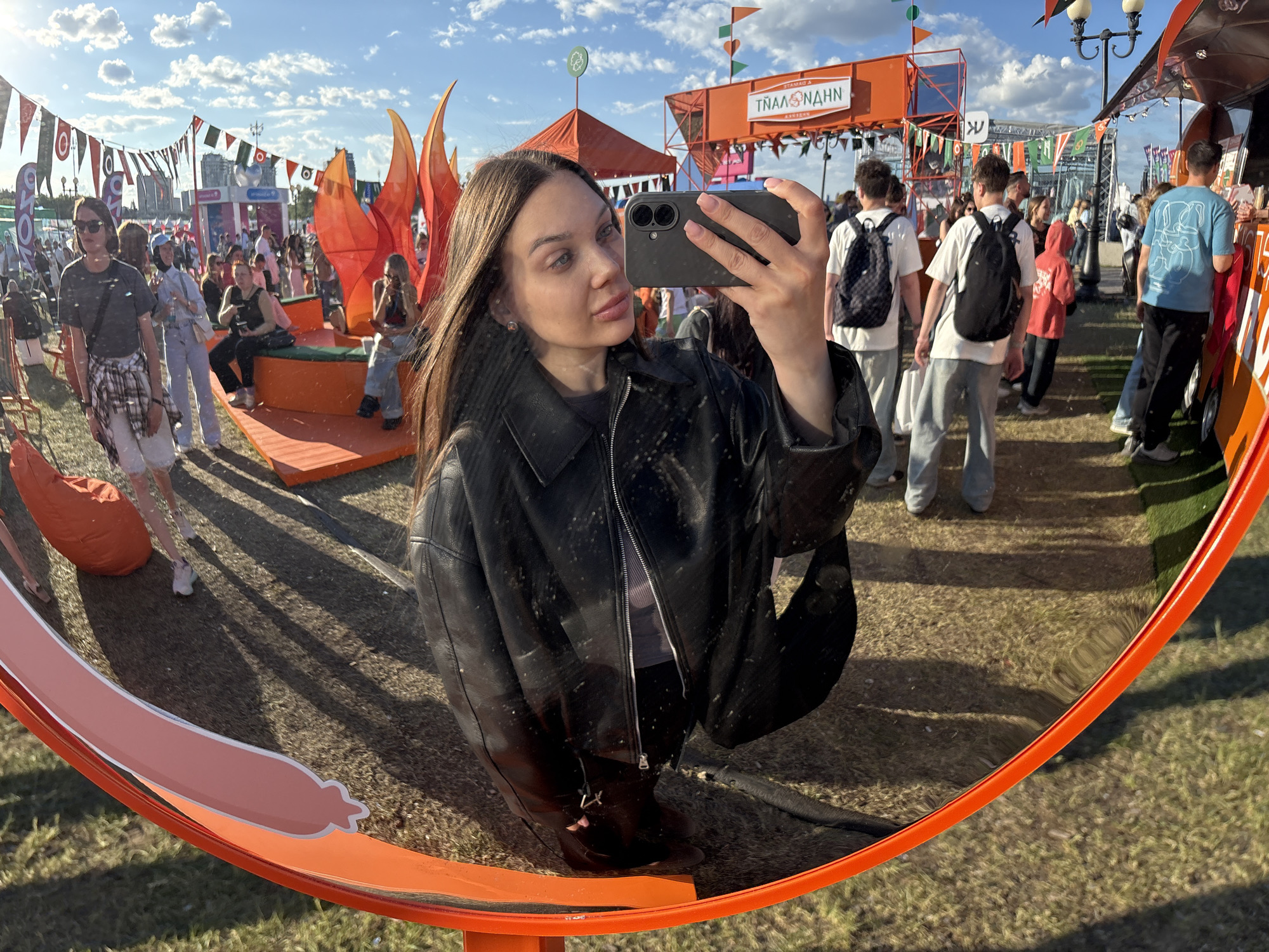 The image depicts a young woman taking a selfie in a large, circular, convex mirror with an orange frame, set outdoors in the city of Kazan, Russia. The woman, positioned centrally, has long dark brown hair and light eyes, wearing a black leather jacket over a grey top. She holds a dark-colored smartphone with a dual-lens camera module, capturing her reflection.
The mirror's reflection reveals an outdoor festival or fair under a bright, partly cloudy blue sky. The ground is covered with sparse grass and patches of darker material, crisscrossed by long shadows from people and objects, indicating strong sunlight from a low angle.
In the reflected scene, numerous individuals are visible throughout the festival area. To the left, a woman in sunglasses, a dark t-shirt, light shorts, and a plaid shirt tied around her waist walks past. Further left, an orange beanbag chair sits on the ground. In the mid-ground, several people are seated on modular orange structures, possibly stylized flame shapes, with one person wearing a light blue jumpsuit. Towards the right, groups of people, predominantly young adults, walk through the area. Several individuals are seen from behind, including two men wearing black backpacks (one with a checkered pattern).
The background features various temporary festival structures, including multiple colorful pennant flags and banners strung across the space. An orange tent or stage-like structure displays Cyrillic text, which appears as "ТЯПЛАНАНН" in the reflection. To the far right, an orange food truck or stall is visible, with a person in a light blue t-shirt standing nearby. In the distant background, a modern city skyline with tall buildings is discernable beneath the horizon. Overhead, white cumulus clouds are scattered across the blue sky. The overall scene conveys a lively, casual outdoor event.