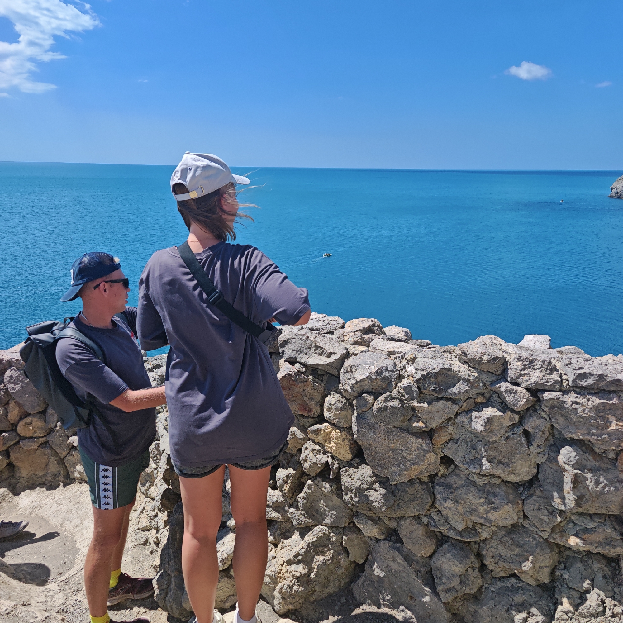 Two individuals are standing on a rocky outcrop overlooking a bright blue sea under a clear sky. The person on the left is a man wearing a dark t-shirt, dark shorts with a white stripe pattern, a dark baseball cap, sunglasses, and a backpack. The person on the right is a woman wearing a dark t-shirt, a light-colored baseball cap, and a dark crossbody bag. They are both facing away from the camera, gazing out at the water. In the distance, a small boat or jet ski is visible on the sea. The environment appears to be a coastal area with rugged terrain and a stone wall in the foreground. This location is the coast of Sudak, Ukraine, known for its scenic sea views.