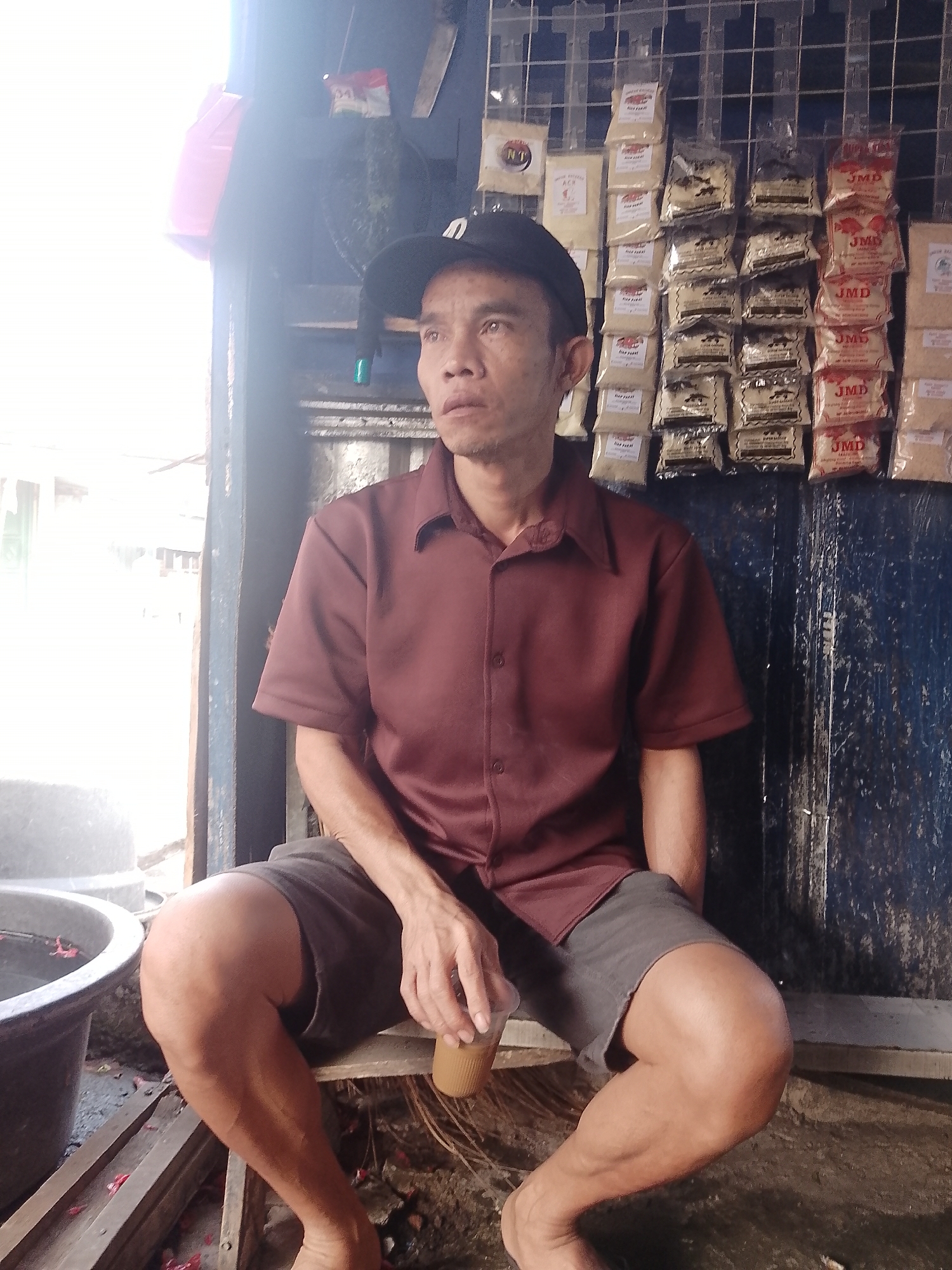 A middle-aged Asian man wearing a dark baseball cap and a maroon collared shirt is seated in front of a display of packaged goods. He is holding a clear plastic cup with a tan liquid in his right hand. The background shows a grid wall with many small, bagged items. To the left of the man is a large, dark gray basin containing water and some pink flower petals. The ground is dirt and debris. The scene appears to be an outdoor market stall in Cikalongkulon, Indonesia.