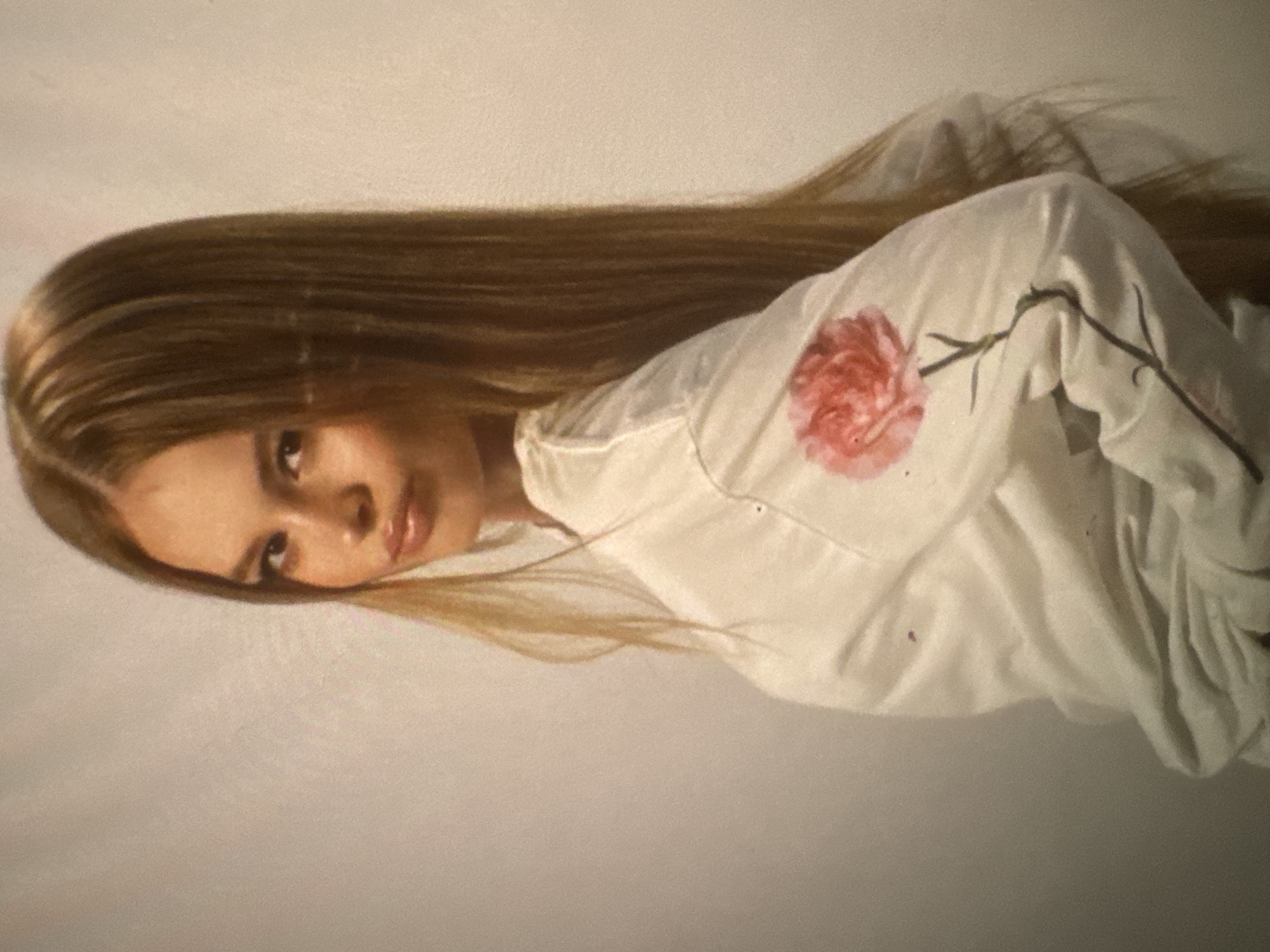 A portrait photograph shows a young woman posing against a plain, light-colored background, likely an off-white or beige wall or studio backdrop. The image appears to be rotated, with the woman's head positioned on the left side of the frame and her body extending to the right.
The woman has very long, straight, light brown hair, which is fanned out to her left, covering part of the background. She looks directly towards the viewer with a calm and gentle expression, her lips slightly parted. Her facial features are soft, and her eyes are open and engaging.
She is wearing a simple, white long-sleeved shirt. A prominent detail on the shirt is a painted or embroidered red/pink flower, resembling a carnation, with a green stem and leaves. This floral design is located near her right shoulder/chest area (as viewed in the rotated image). The fabric of the shirt shows some natural folds and a few small, dark specks.
The setting is minimalist, providing no specific location cues beyond suggesting an indoor or controlled environment. The lighting is soft and even, highlighting her features and hair without harsh shadows. There is no visible text in the image. The overall impression is one of serene beauty and simplicity.