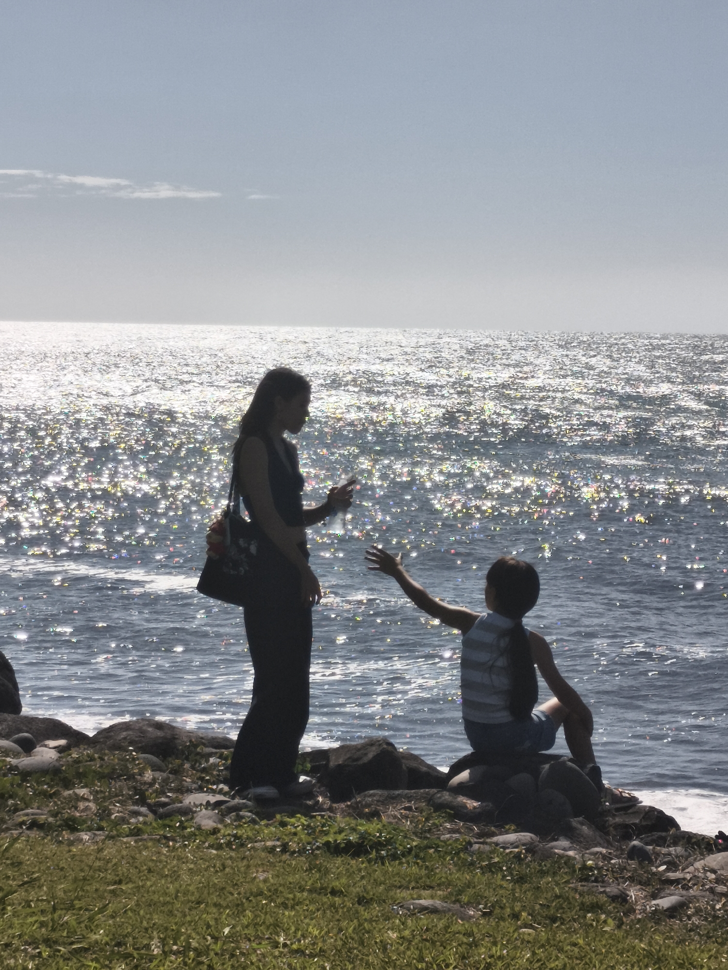 Two silhouetted figures stand on a rocky shoreline next to the sparkling ocean under a hazy sky. A woman on the left, wearing dark clothing and carrying a bag, holds a bottle in her right hand. A child on the right, wearing a striped shirt and shorts, sits on rocks facing the ocean with their right arm extended. The location is the city of Sainte-Marie, France, with the sun reflecting intensely off the water's surface.