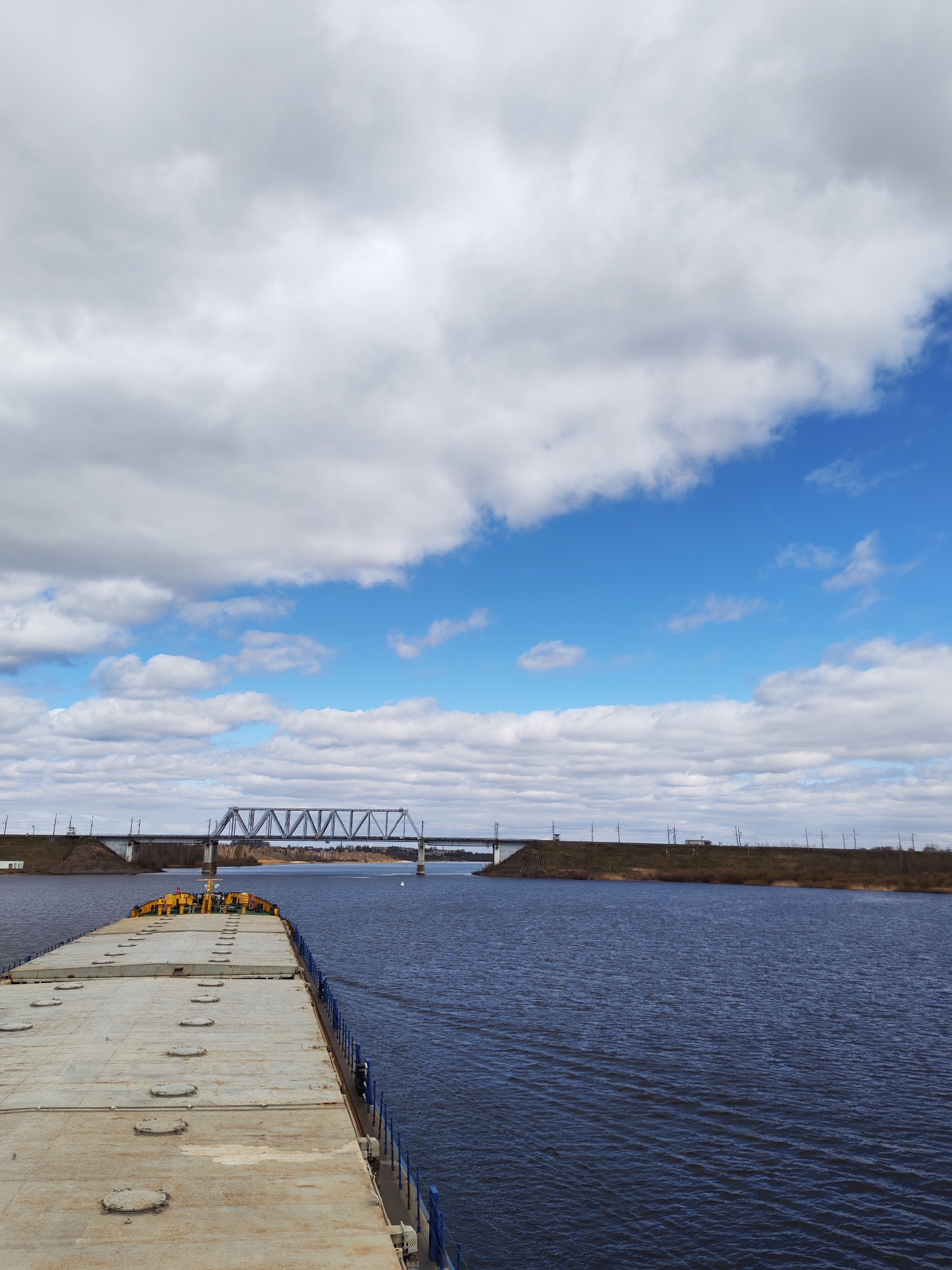 The image depicts a wide river with a long, flat barge in the foreground. The barge has a dull gray surface with numerous circular hatches and a low blue railing along its edge. In the background, a steel truss bridge spans the river, connecting to land on either side. The riverbanks are vegetated, with patches of brown grass and sparse trees. The sky above is blue, filled with puffy white clouds. The location and city are unknown.