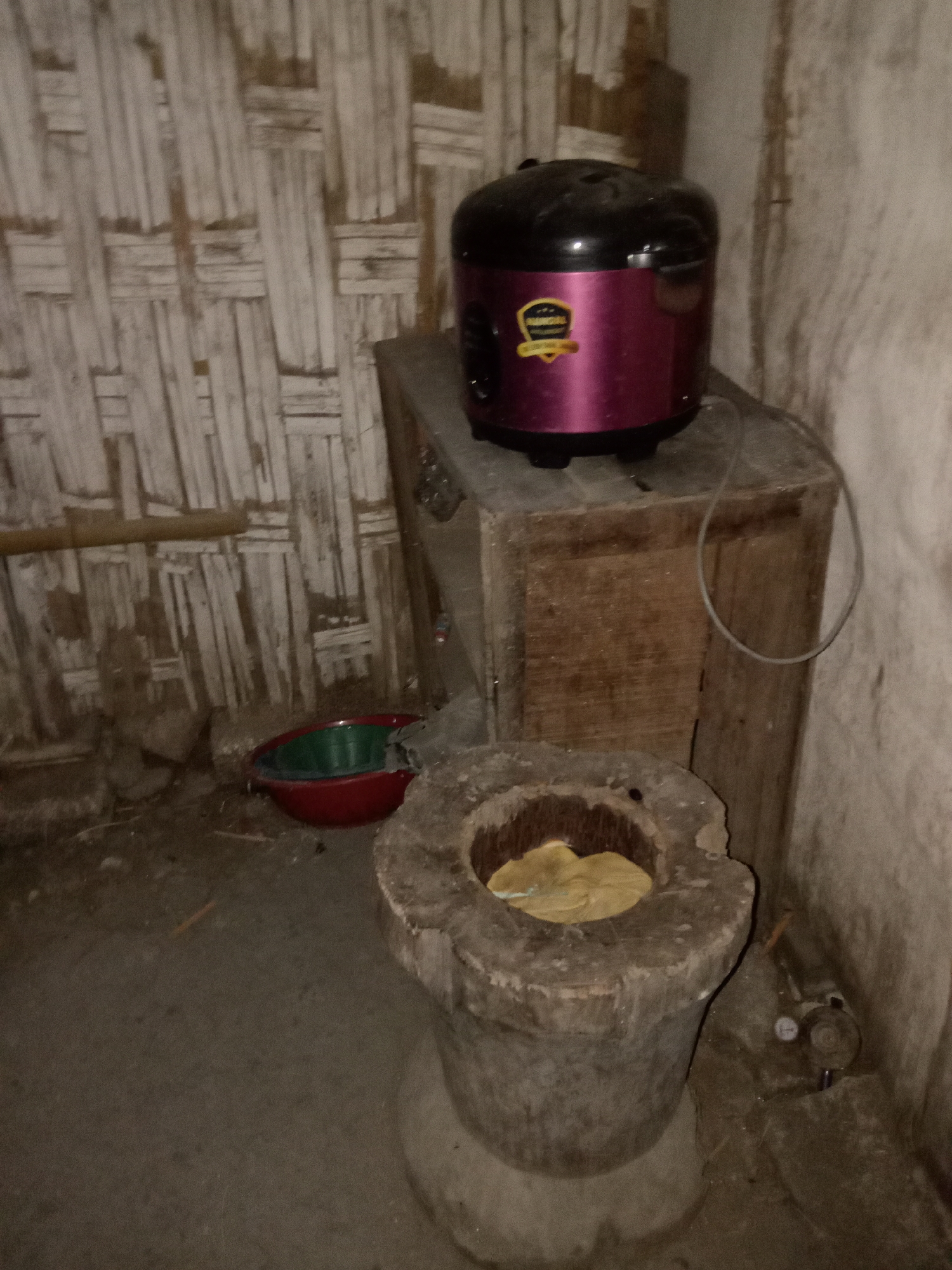 This image depicts an indoor scene, likely a kitchen or utility area, located in Jumput, Indonesia. The walls are constructed from woven bamboo or reeds, a common building material in the region. In the center of the frame, a wooden cabinet stands against the wall, topped with a small, burgundy and black rice cooker. To the left of the cabinet, on the floor, rests a red and green plastic bowl. In the foreground, a large, wooden mortar, a traditional tool for grinding food, is visible. The floor is made of concrete. The lighting in the image is dim, suggesting an evening or indoor setting with limited natural light. There are no people present in the scene. The overall impression is one of a humble, functional living space.
