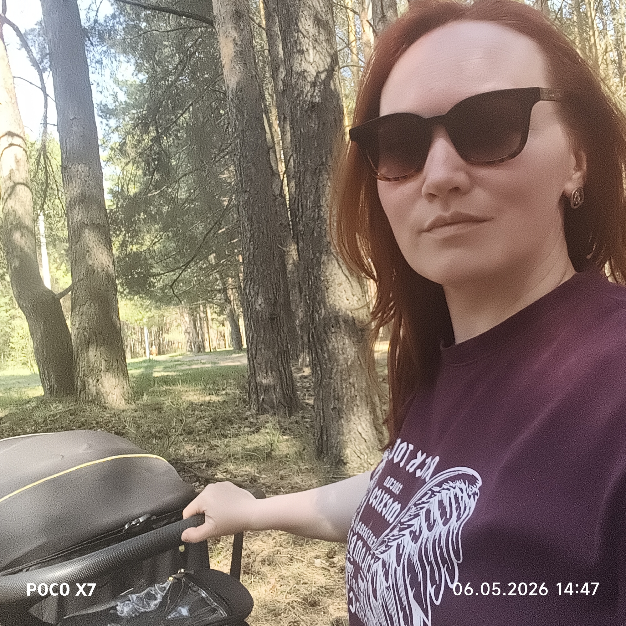 A woman with auburn hair and sunglasses is in a forest in the city of Chaikovsky, Russia. She is wearing a dark purple t-shirt and holding the handle of a stroller. Several tall trees surround the stroller. The time stamp indicates 14:47 on May 6, 2026.