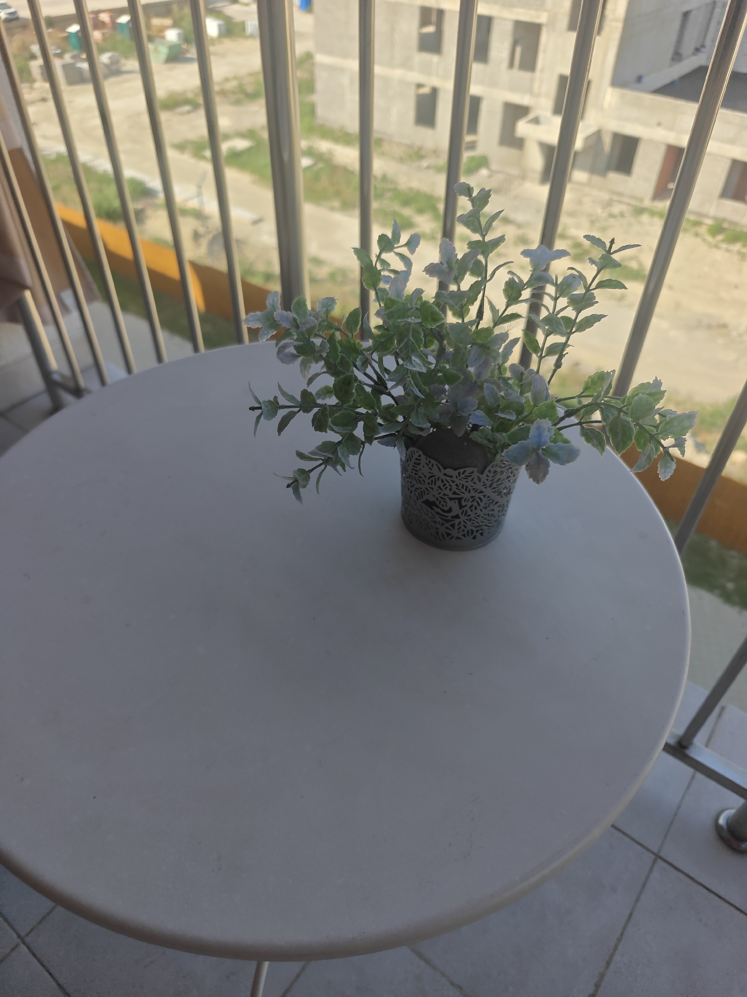 A close-up, high-angle shot features a round, white table on a balcony. A small potted artificial plant with green and bluish-white leaves sits on the right side of the table. The plant is in a decorative, silver-colored pot with an intricate cut-out pattern. In the background, through a series of vertical metal railings, a construction site is visible with unfinished buildings and some portable toilets. The ground outside the balcony appears to be a mix of dirt and sparse green grass. The scene is bathed in warm, natural light, suggesting it is daytime.