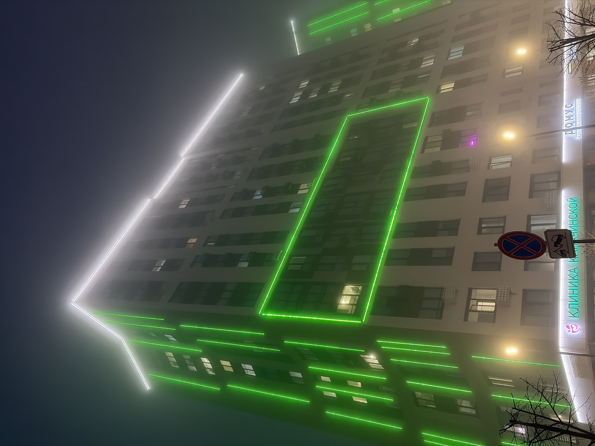 At night, a multi-story building is illuminated with green and white lights in Izhevsk, Russia. The building is a pale color, possibly white or light gray. There are rows of windows across its facade. The lighting design includes a prominent white outline that forms an angular shape at the top and extends down the side of the building. Green lights are also arranged in horizontal lines, tracing architectural features, and forming rectangular shapes on the building’s surface. There are streetlights nearby that add to the scene's illumination. To the right of the building, there is signage in Cyrillic, and a traffic sign is partially visible. Trees with bare branches are silhouetted in the foreground. The overall atmosphere is misty or foggy, which softens the light and obscures the details of the surrounding environment.