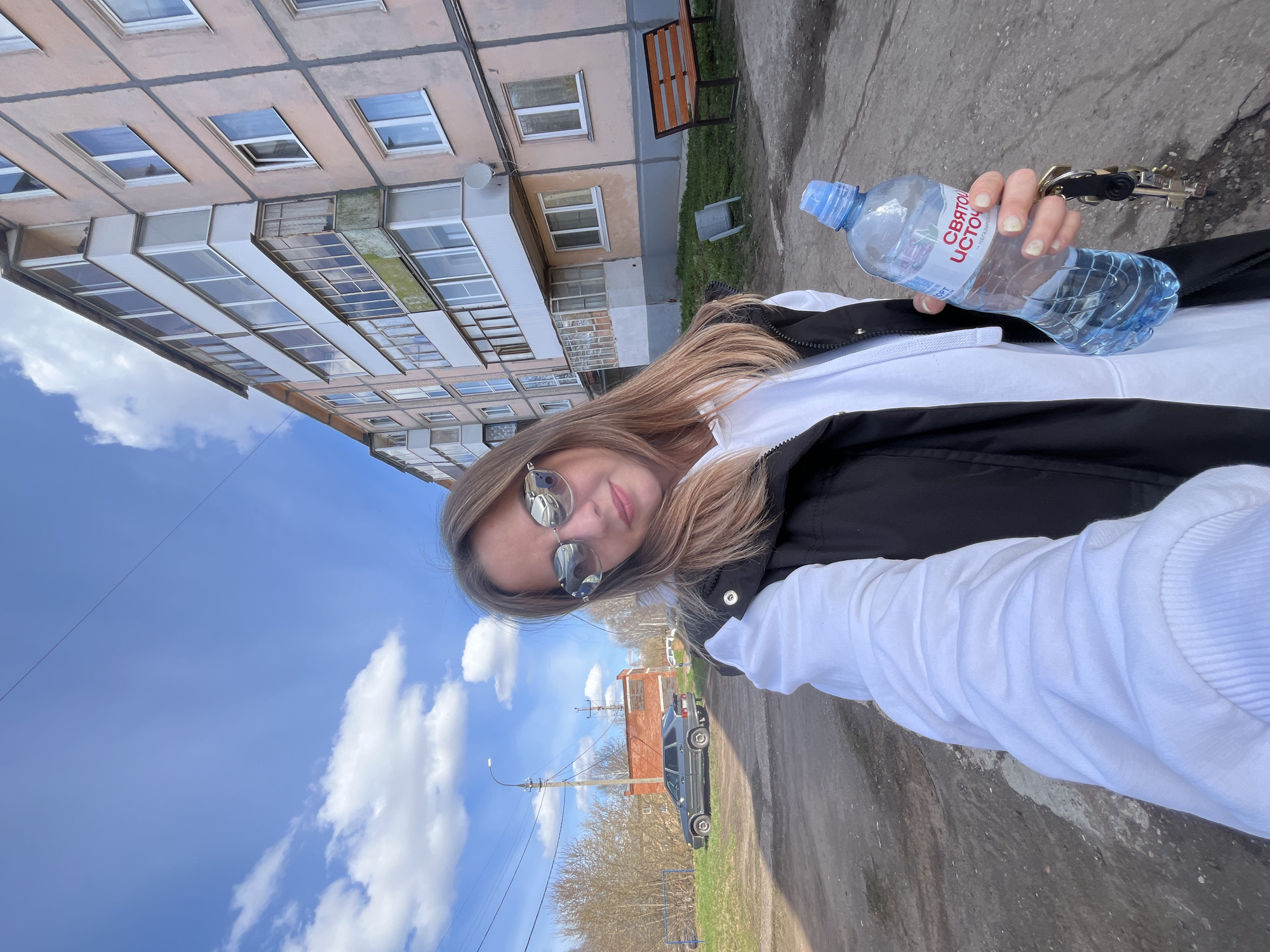 A daytime selfie captured outdoors in Votkinsk, Russia, shows a woman smiling subtly at the camera. She has long, light brown hair and is wearing reflective sunglasses, a white hoodie, and a black vest. In her right hand, she holds a plastic water bottle prominently labeled "СВЯТОЙ ИСТОЧНИК" (Svyatoy Istochnik, meaning Holy Spring) and a set of keys.
The background features a multi-story residential building with a worn, light-pink facade and multiple windows and balconies, positioned vertically in the left portion of the image. A wooden bench is visible near the building's base. To the right, there is a dirt or gravel path, and further back, an older dark-colored car is parked near a red brick building and a utility pole. Bare trees suggest an early spring or late autumn season. The sky is bright blue with several white, fluffy clouds, indicating clear, sunny weather. The image is composed at a tilted angle, giving a dynamic perspective to the scene.