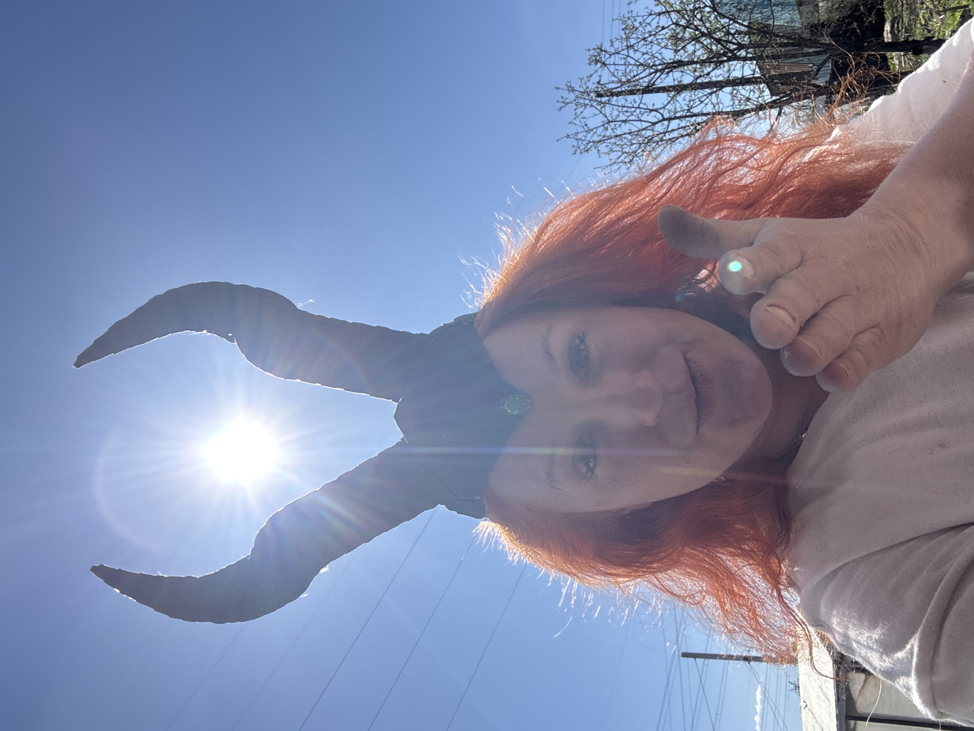 A woman with red hair is outdoors, facing upwards at the camera. She is wearing a set of large, dark-colored, curved horns on her head. Her right hand is raised, giving a thumbs-up. The sky is a clear, bright blue with the sun shining directly behind one of the horns, creating a lens flare effect. The environment includes bare tree branches and what appears to be a glimpse of a building or structure in the background. It is difficult to definitively identify the location as Chaikovsky, Russia from the details provided in the image.