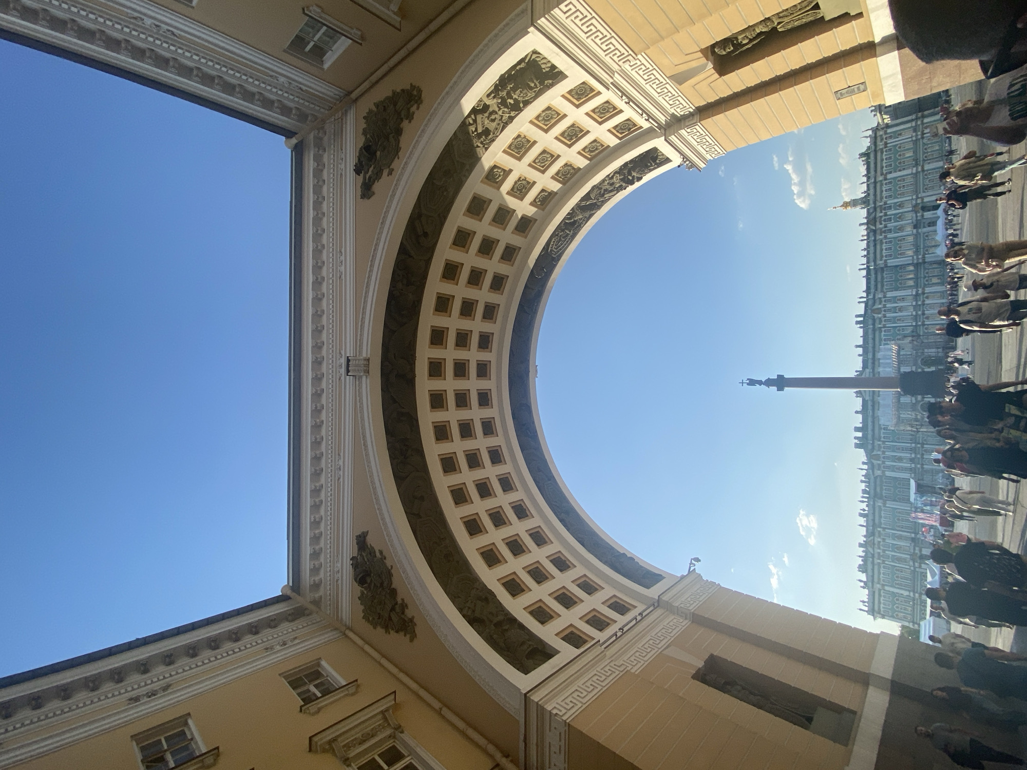 The image shows the General Staff Building's Triumphal Arch in Saint Petersburg, Russia, framing the Palace Square. The archway's interior features a coffered ceiling and bas-relief sculptures. In the background, the Alexander Column and a portion of the Winter Palace are visible. Numerous people are present in the square, suggesting pedestrian traffic and possibly a gathering. The sky is clear and blue.